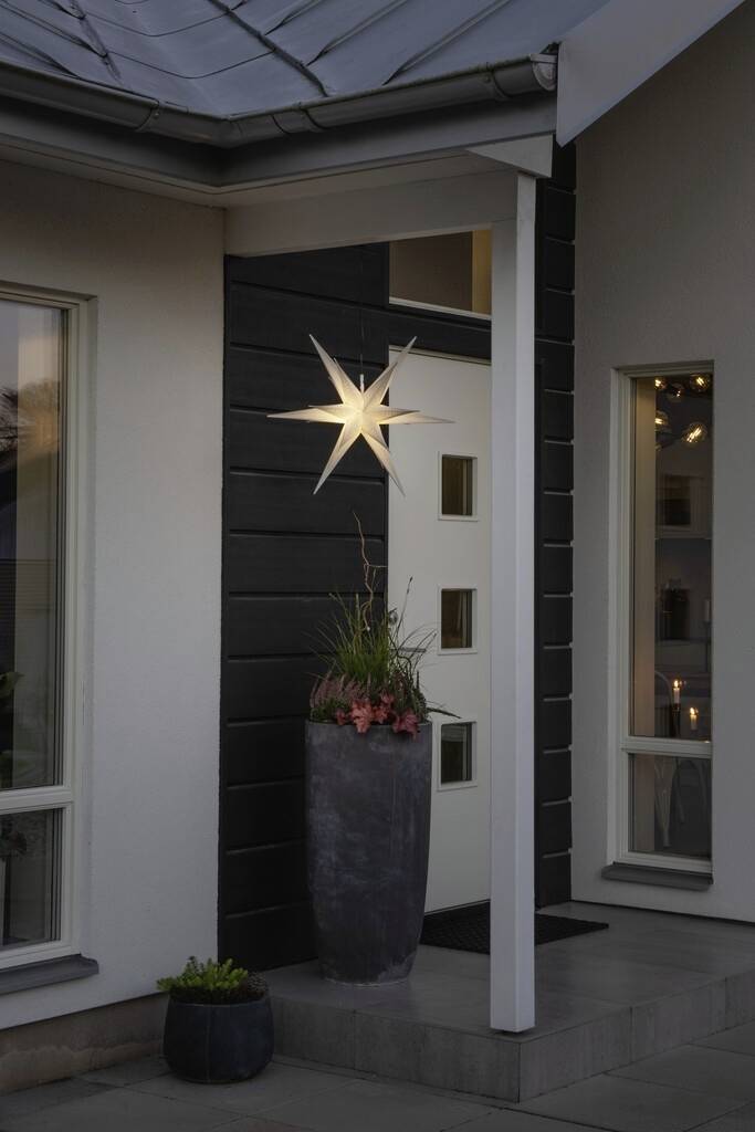 Ein beleuchteter Stern hängt an der Außenwand eines modernen Hauses. Daneben steht ein hoher Pflanzkübel mit Blumen.