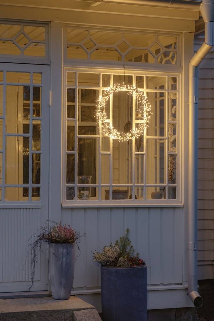 Ein beleuchteter Kranz hängt innen an einem Fenster eines Hauses mit dekorativen Rahmen. Draußen stehen zwei Pflanzgefäße auf der Veranda.