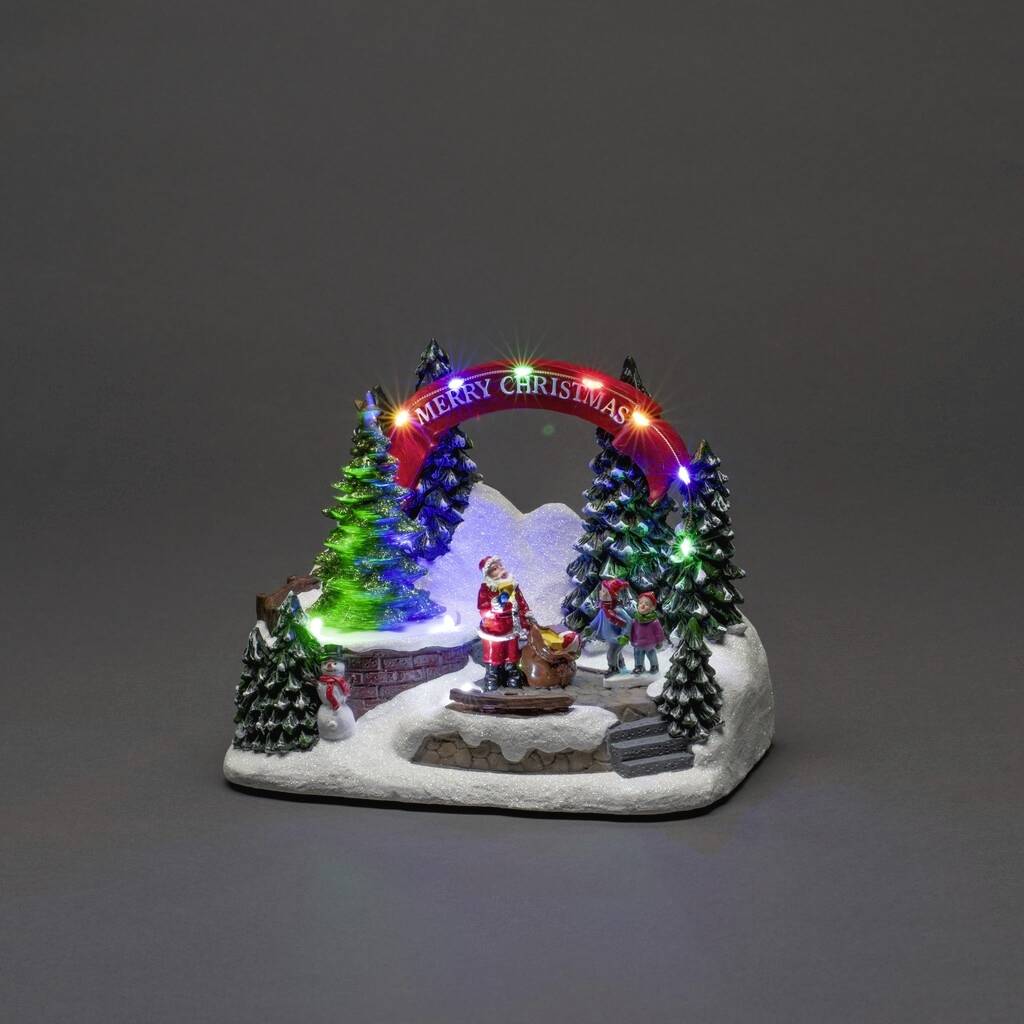 Weihnachtliches Diorama mit beleuchteten Tannenbäumen, einem Weihnachtsmann mit Schlitten und einem leuchtenden 'Merry Christmas'-Bogen.