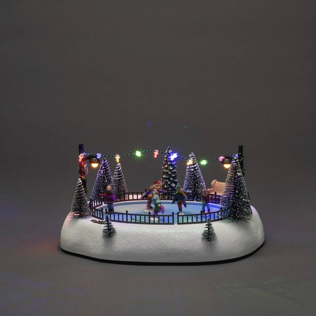 Ein Minatur-Weihnachtsdorf mit Eislaufbahn. Figuren eislaufen zwischen beleuchteten Tannen. Dekorative Lichterketten um den Rand.