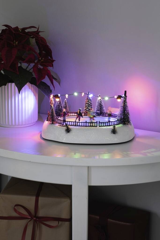 Miniatur-Schlittschuhlaufbahn auf Tisch, umgeben von kleinen beleuchteten Tannenbäumen, mit einem Poinsettia-Topf daneben.
