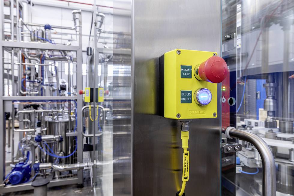 Gelber Not-Aus-Knopf an einer industriellen Maschine, umgeben von Metallleitungen und Geräten in einer Fabrikumgebung.