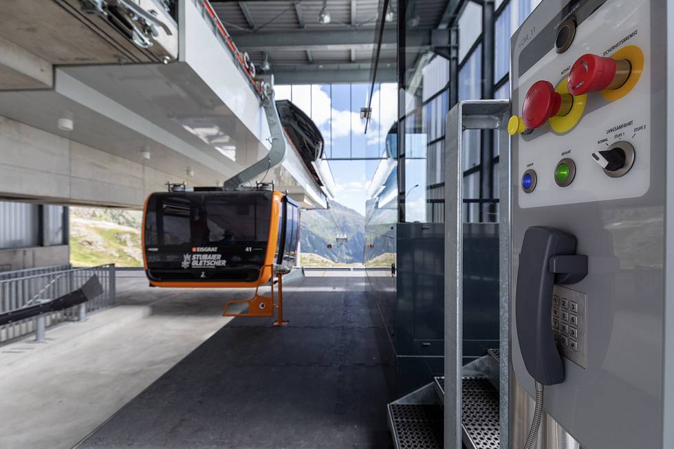 Seilbahnstation mit leerer Gondel, Berglandschaft im Hintergrund, Bedienfeld mit Knöpfen und Telefon im Vordergrund.