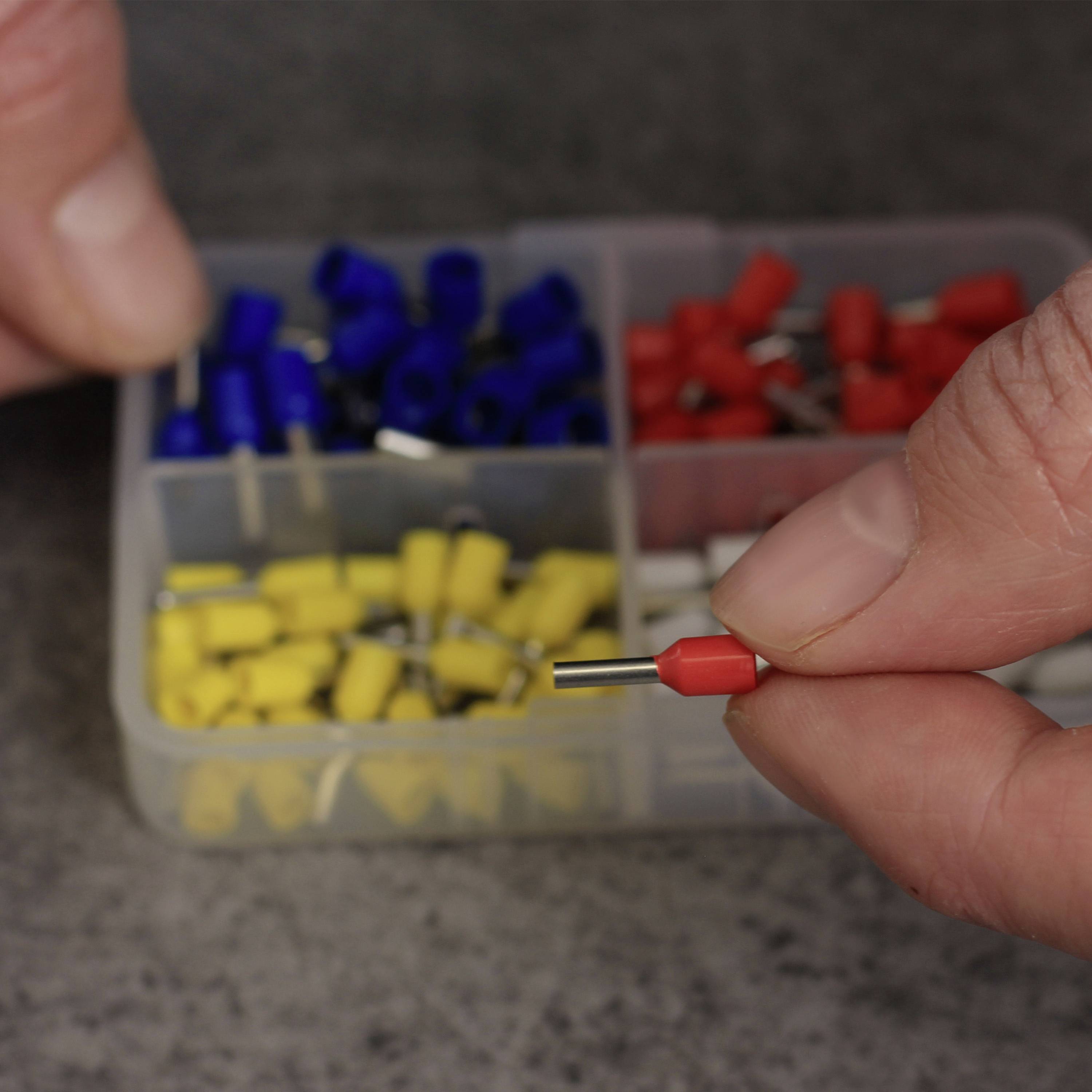 Zwei Hände, eine hält einen gelben Kabelstecker, während die andere einen roten in einer Plastikkiste voller farbiger Stecker zeigt.