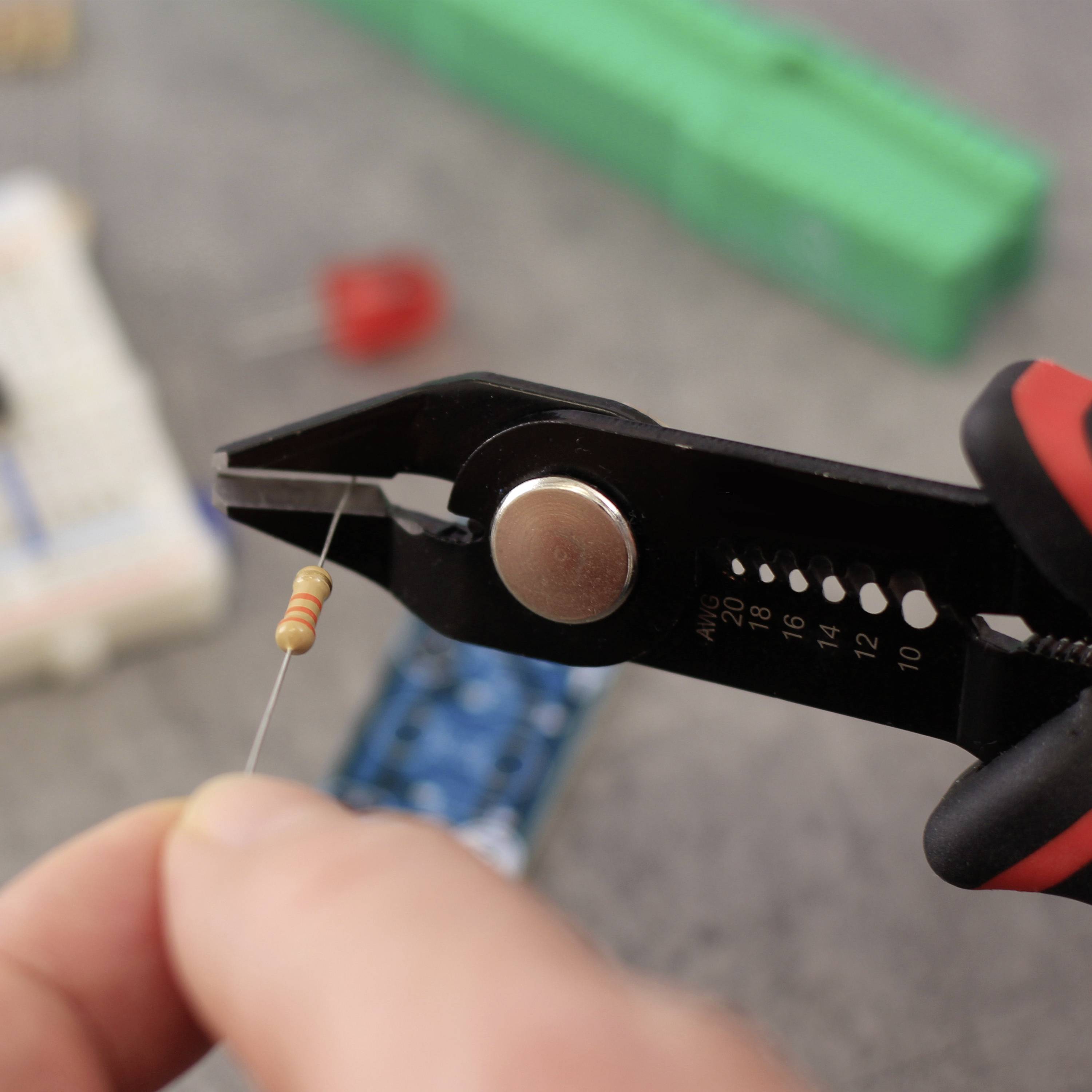 Eine Person hält eine Zange und kürzt einen Widerstandsdraht, umgeben von elektronischen Bauteilen auf einem Tisch.