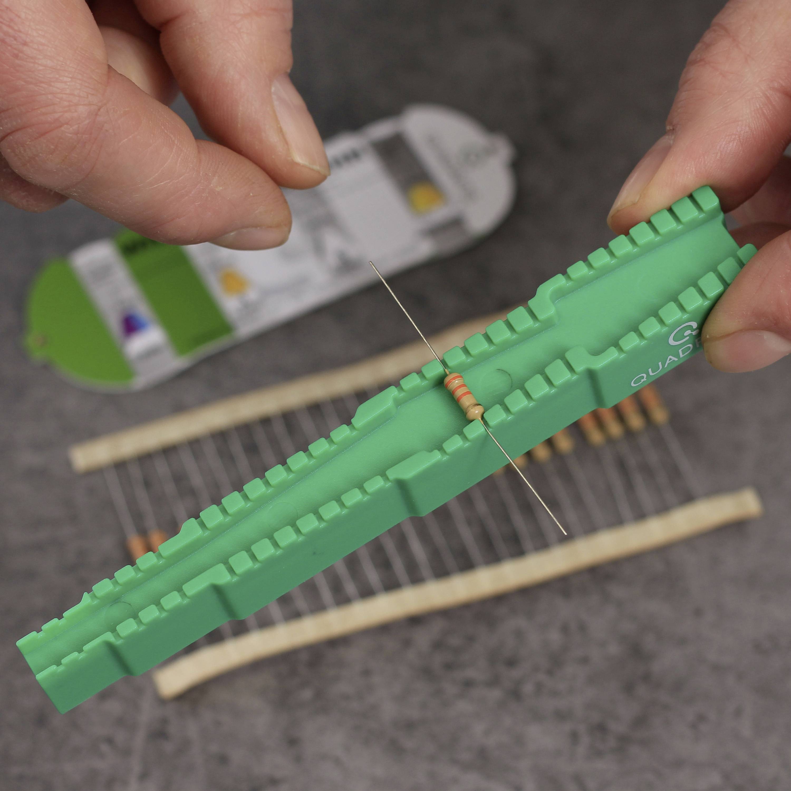 Eine Hand hält einen weißen Widerstandsprüfer mit orangem Widerstand, im Hintergrund liegen weitere Widerstände auf einem Tisch.