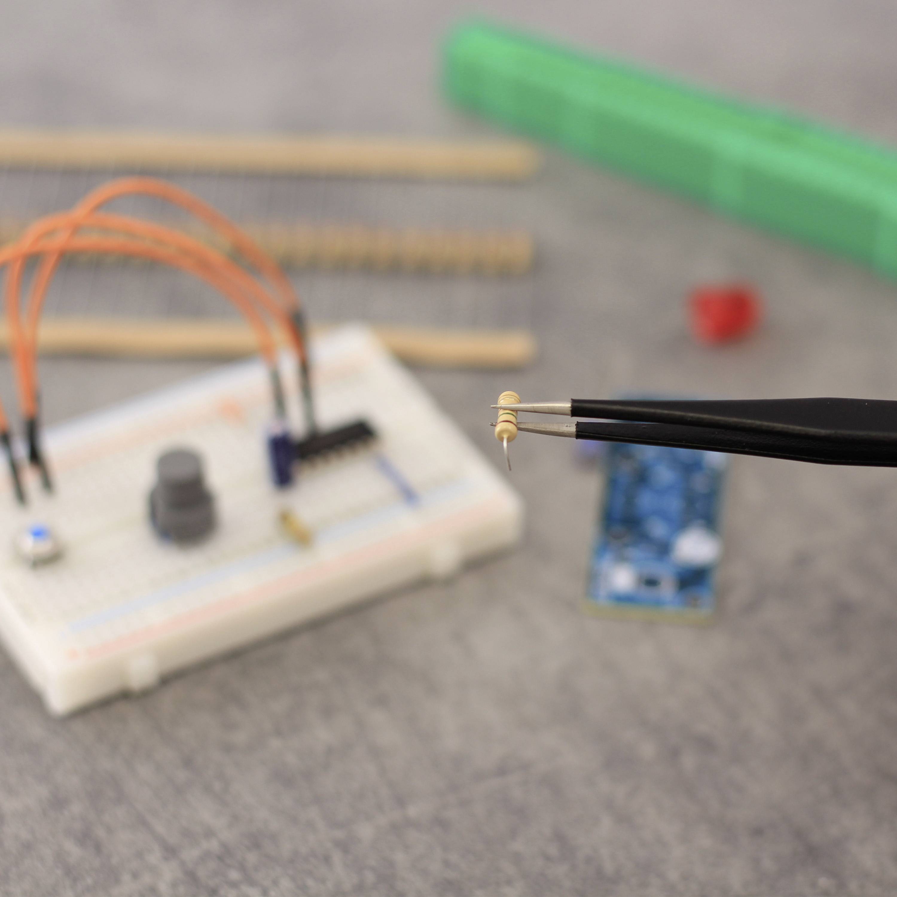 Eine Hand hält mit einer Pinzette einen kleinen elektrischen Widerstand über einem Experimentierboard mit Kabeln und Schaltungen auf einem Tisch.