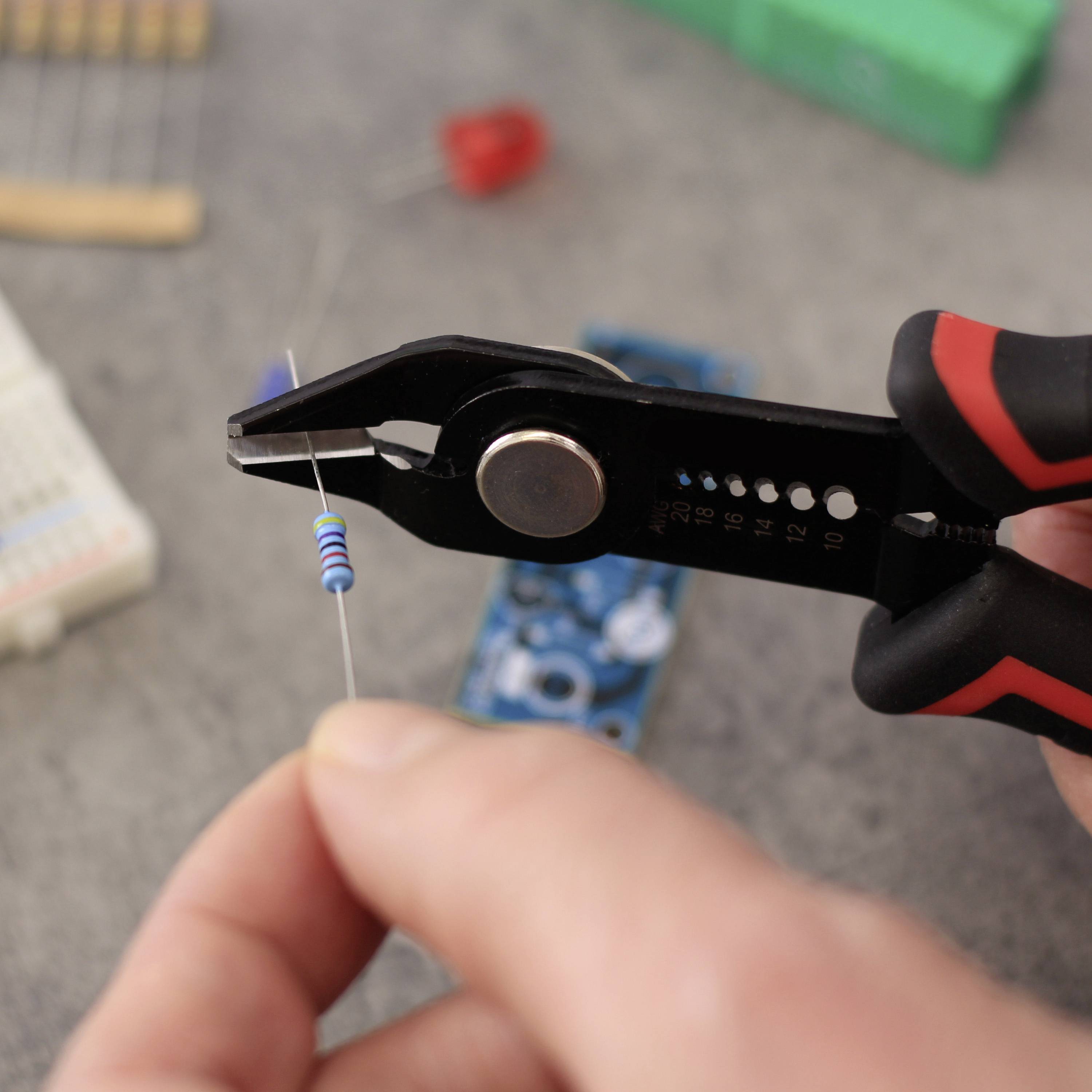 Eine Hand hält eine Zange, die einen kleinen blauen Widerstand für elektronische Schaltungen verbiegt. Im Hintergrund Werkbankzubehör.