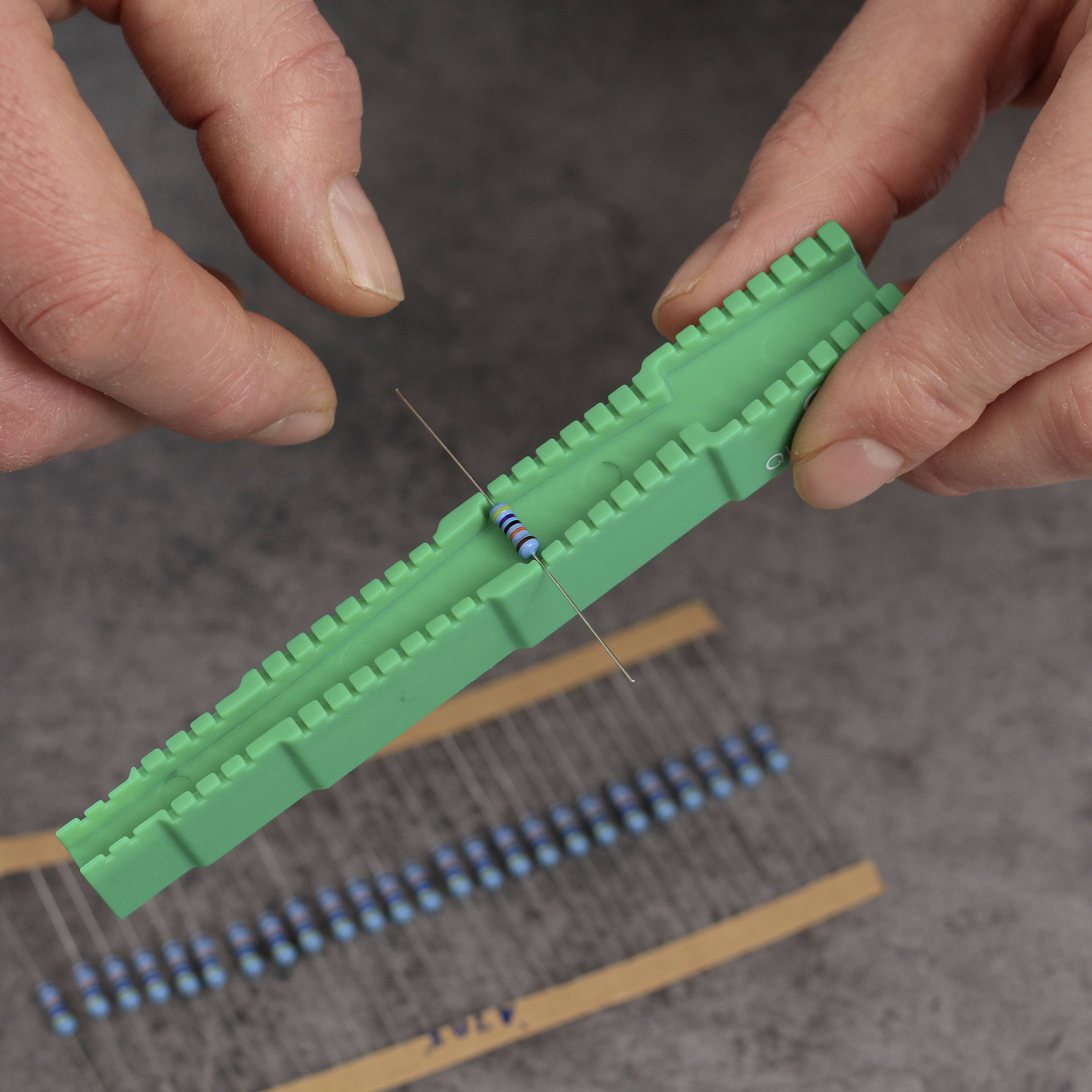 Zwei Hände halten einen grünen Widerstandshalter mit einem blauen axialen Widerstand. Weitere Widerstände liegen im Hintergrund.