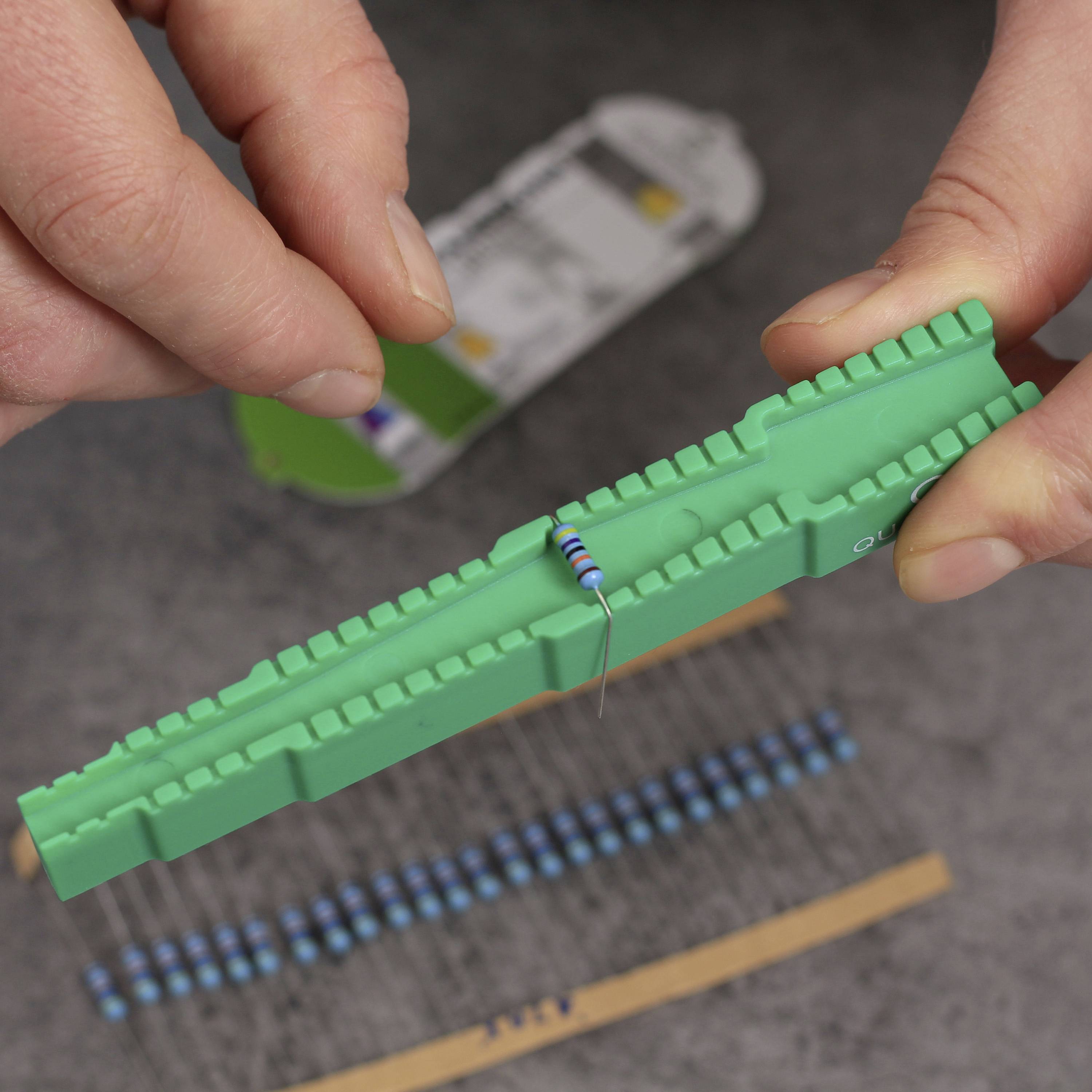 Zwei Hände halten einen grünen Speicherriegel mit einem blauen Widerstand darauf. Im Hintergrund ist unscharf ein weiteres elektrisches Bauteil zu sehen.