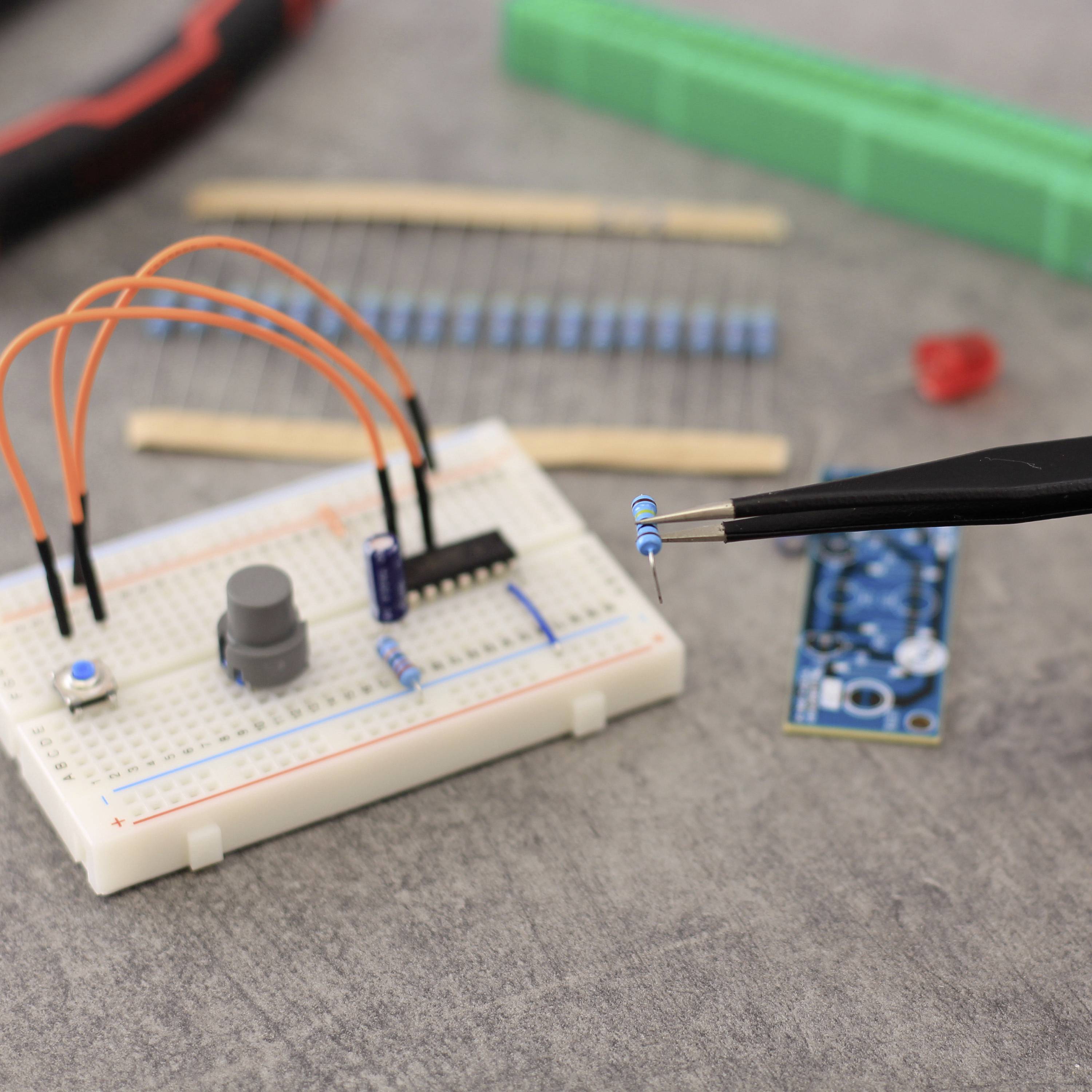 Elektronik-Arbeitsplatz mit Steckplatine, Widerständen, Kabeln und Werkzeug. Eine Person hält einen Widerstand mit einer Pinzette.