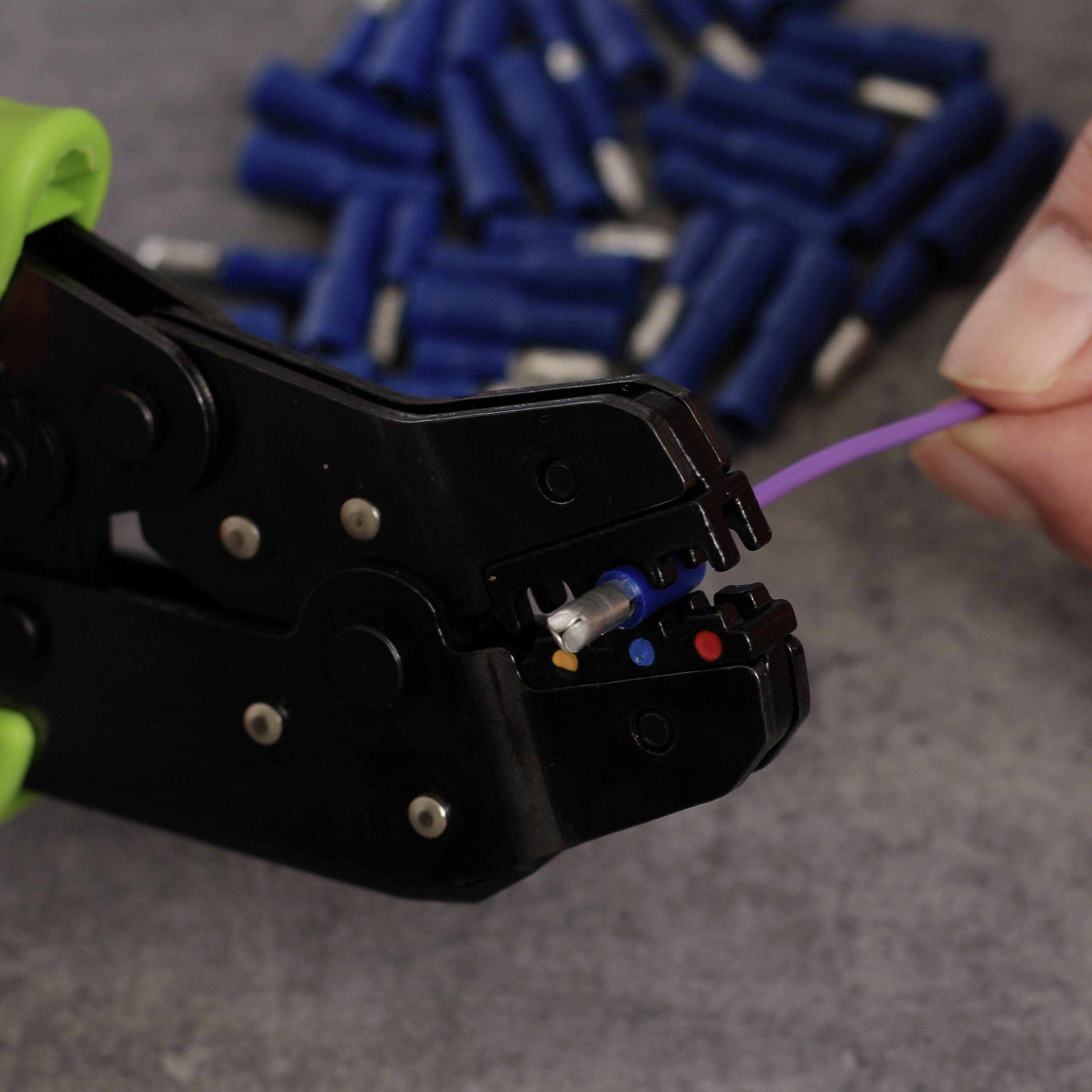 Eine Crimpzange verbindet ein lila Kabel mit einem blauen Kabelschuh. Im Hintergrund liegen blaue Kabelschuhe.