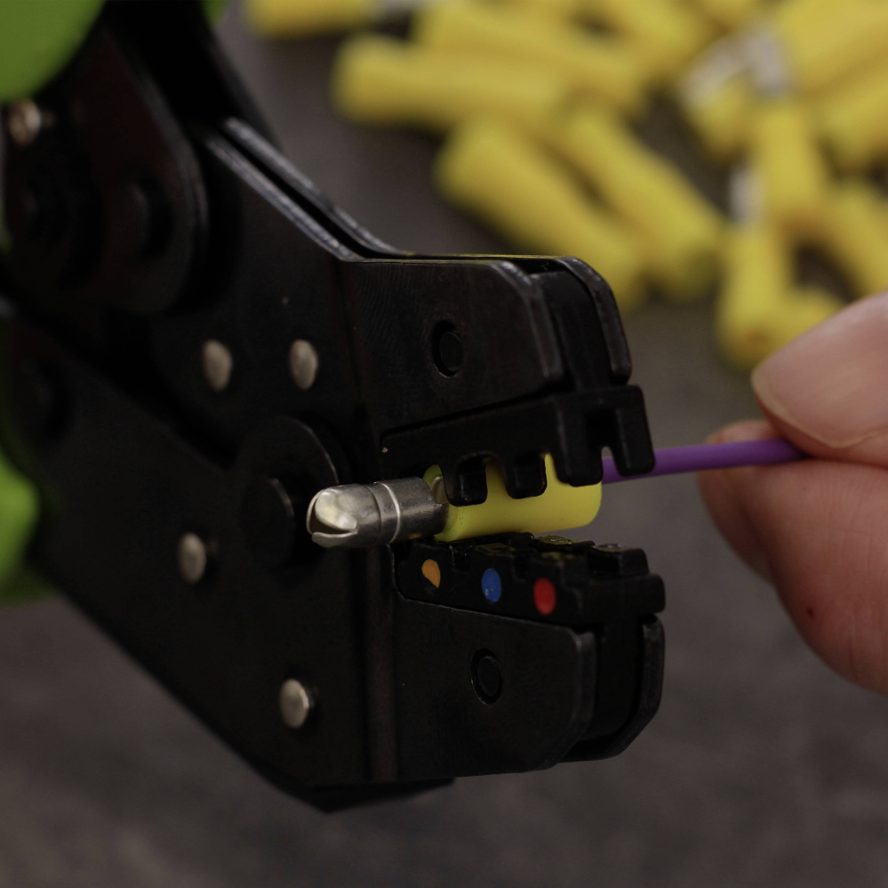 Eine Hand verwendet ein Crimpwerkzeug, um ein lila Kabel mit einem gelben Kabelschuh zu verbinden. Im Hintergrund sind unscharfe gelbe Teile zu sehen.