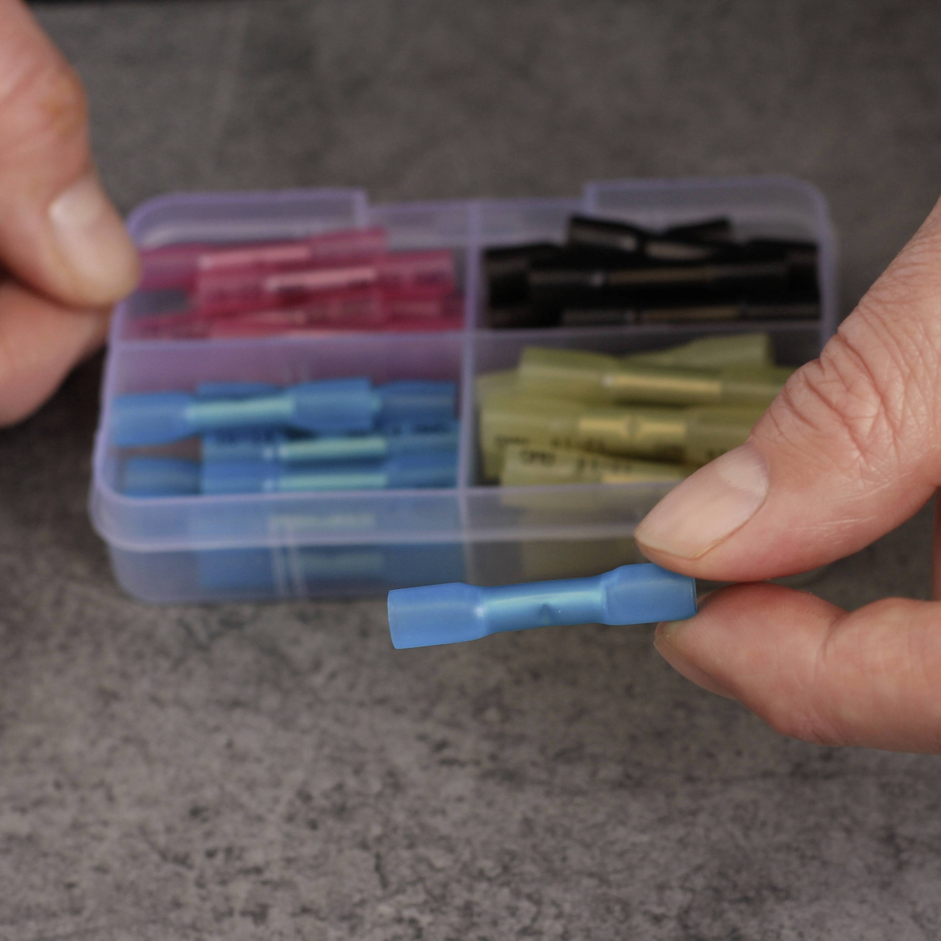 Zwei Hände halten einen blauen Kabelverbinder über einem Tisch. Dahinter ist eine geöffnete Box mit verschiedenen farbigen Verbindern.