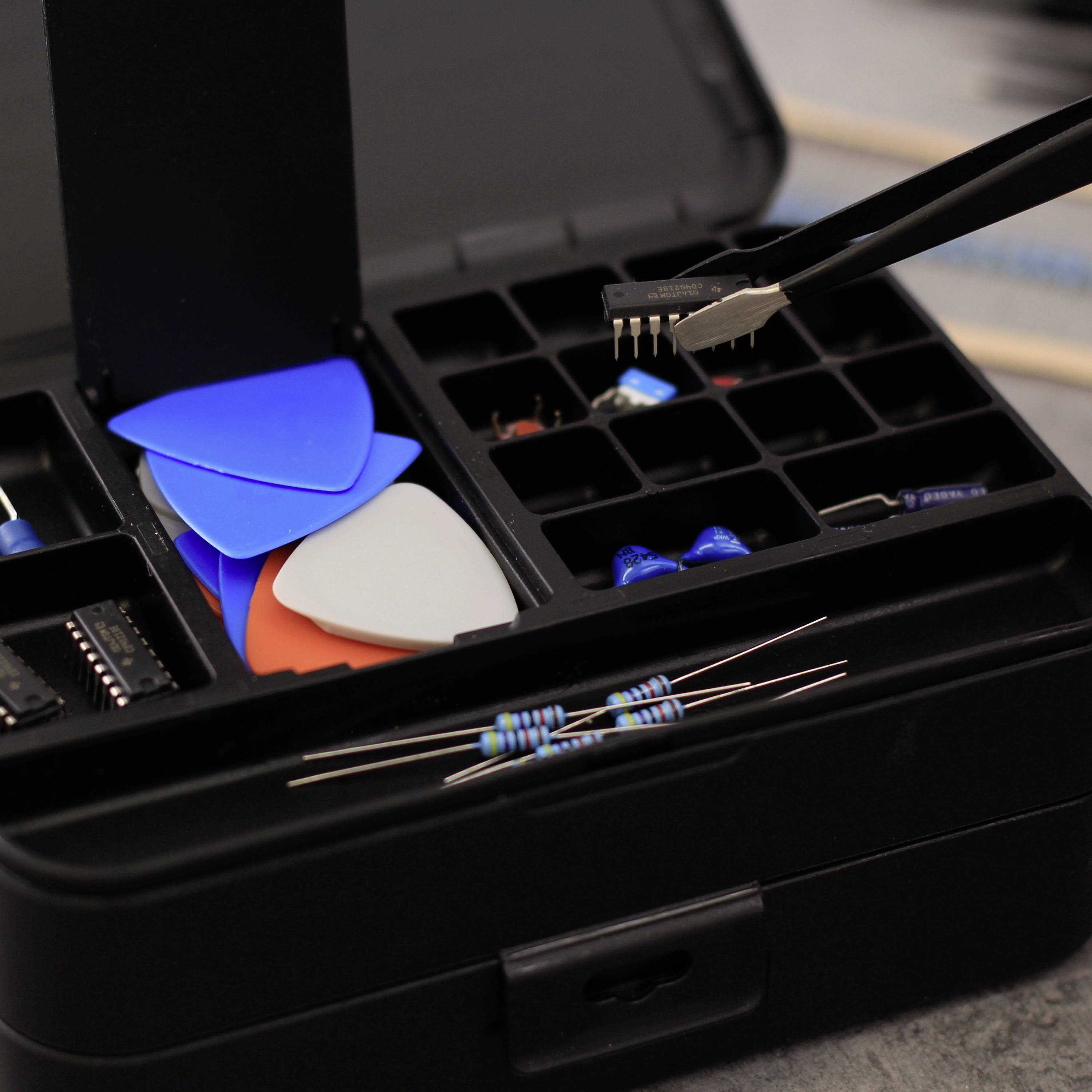 Ein offener Werkzeugkasten mit Gitarrenplektren, Elektronikteilen wie Widerständen und Mikrochip. Ein Werkzeug hält einen Mikrochip über dem Kasten.