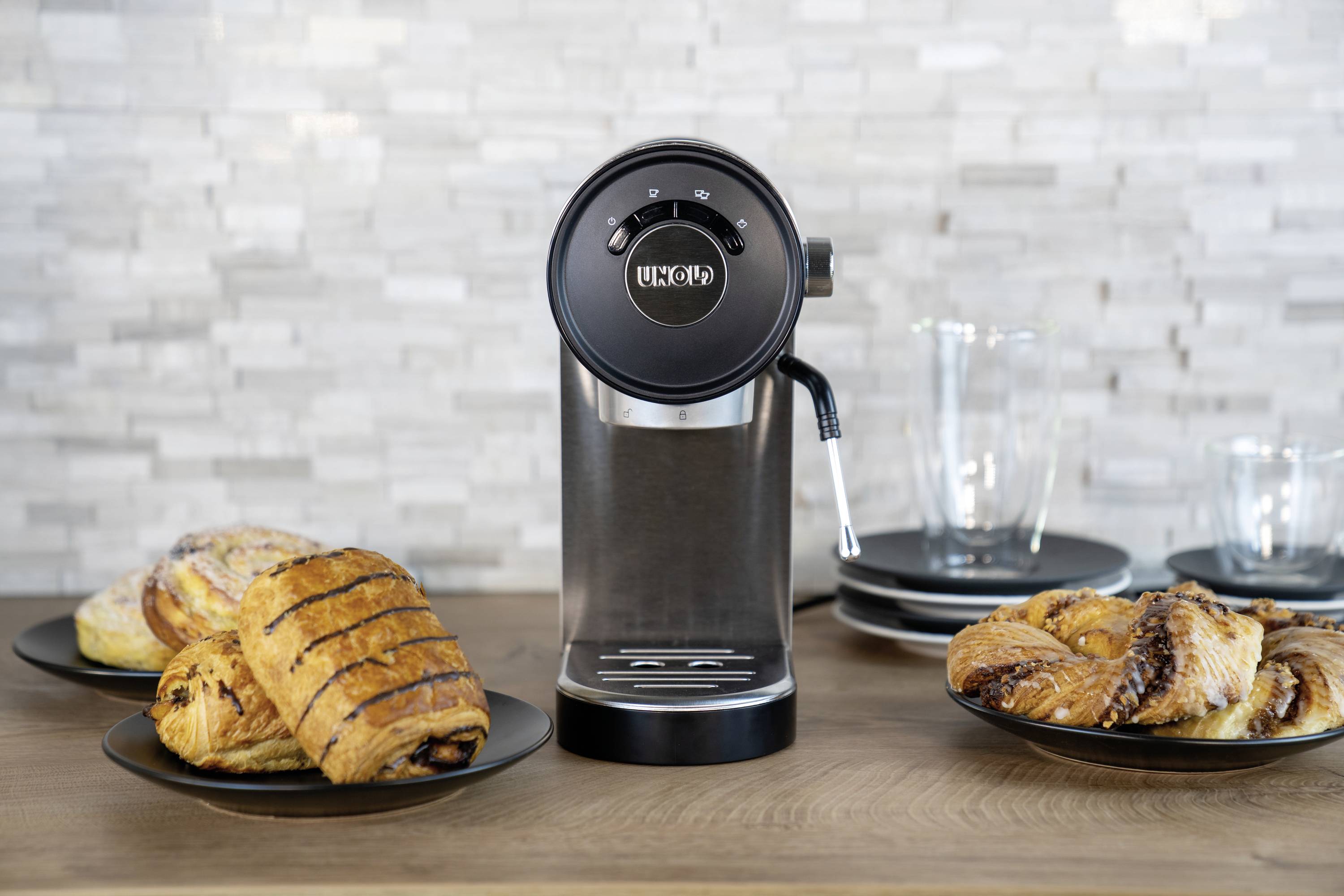 Eine schwarze Kaffeemaschine steht auf einem Holztisch, umgeben von Tellern mit frisch gebackenen Croissants und Gebäck.