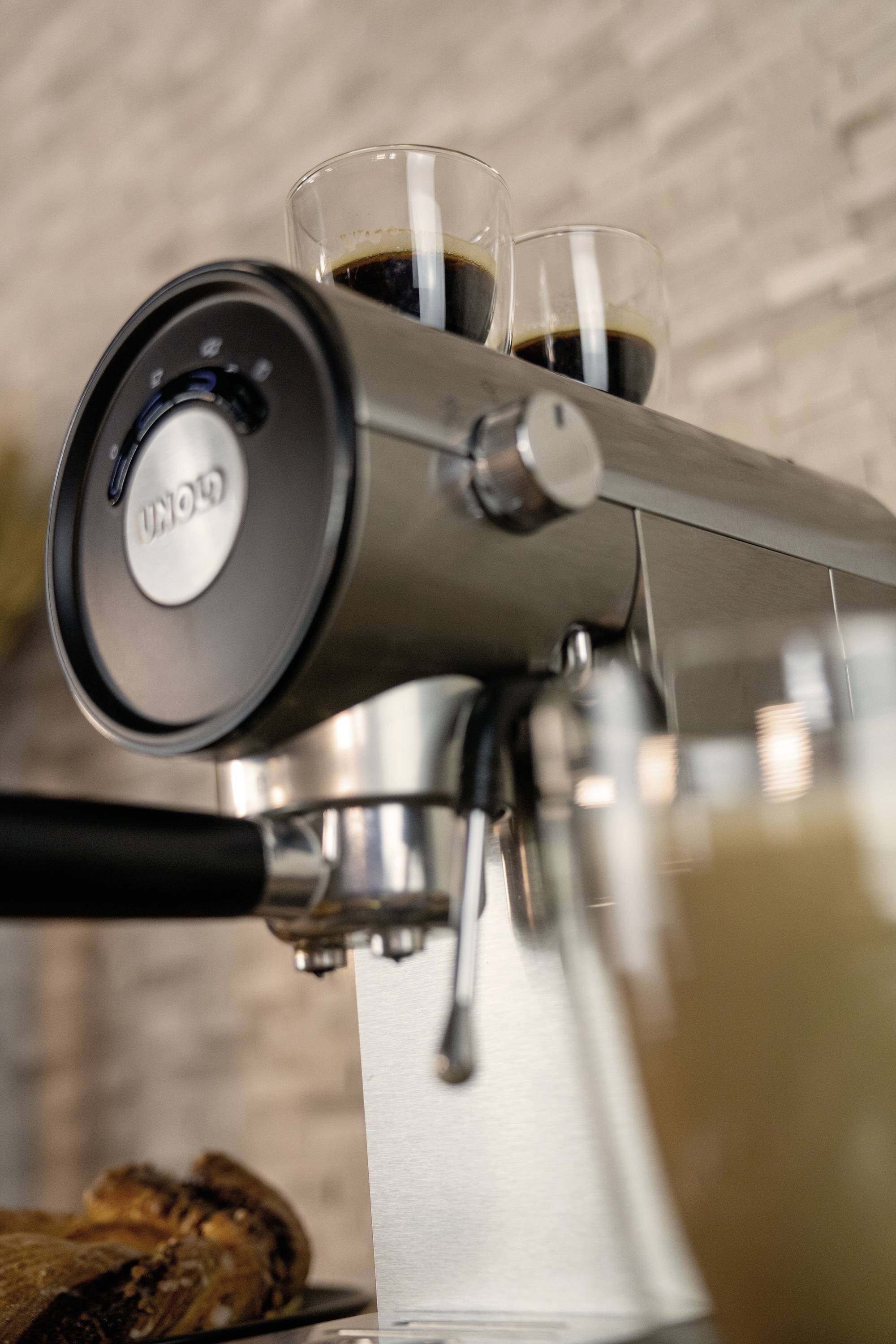 Espressomaschine mit zwei Gläsern Espresso oben, im Hintergrund verschwommenes Café-Setting.