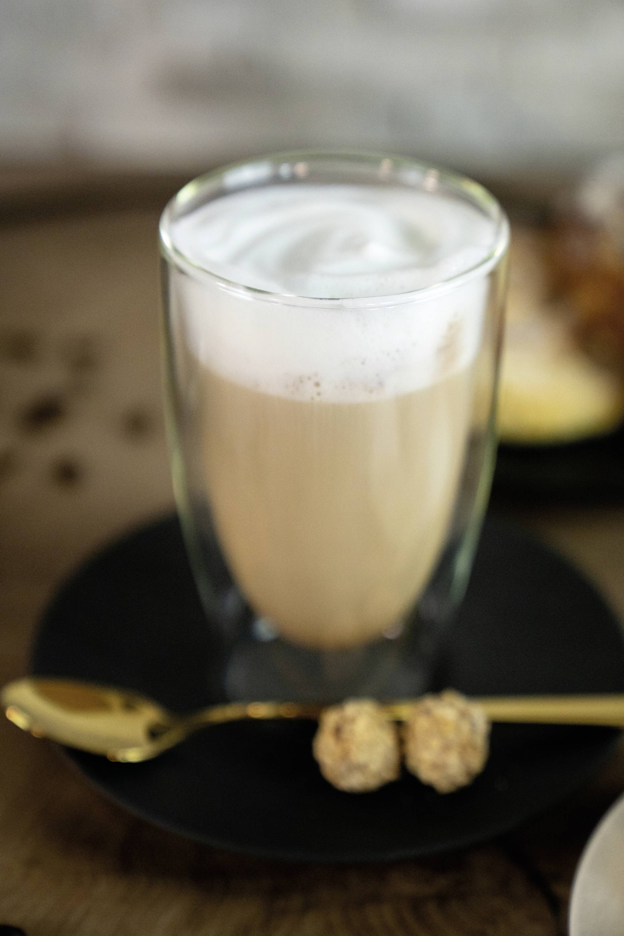 Ein Glas Kaffeegetränk mit cremigem Milchschaum, serviert auf einem Teller mit zwei Pralinen auf einem goldenen Löffel daneben.