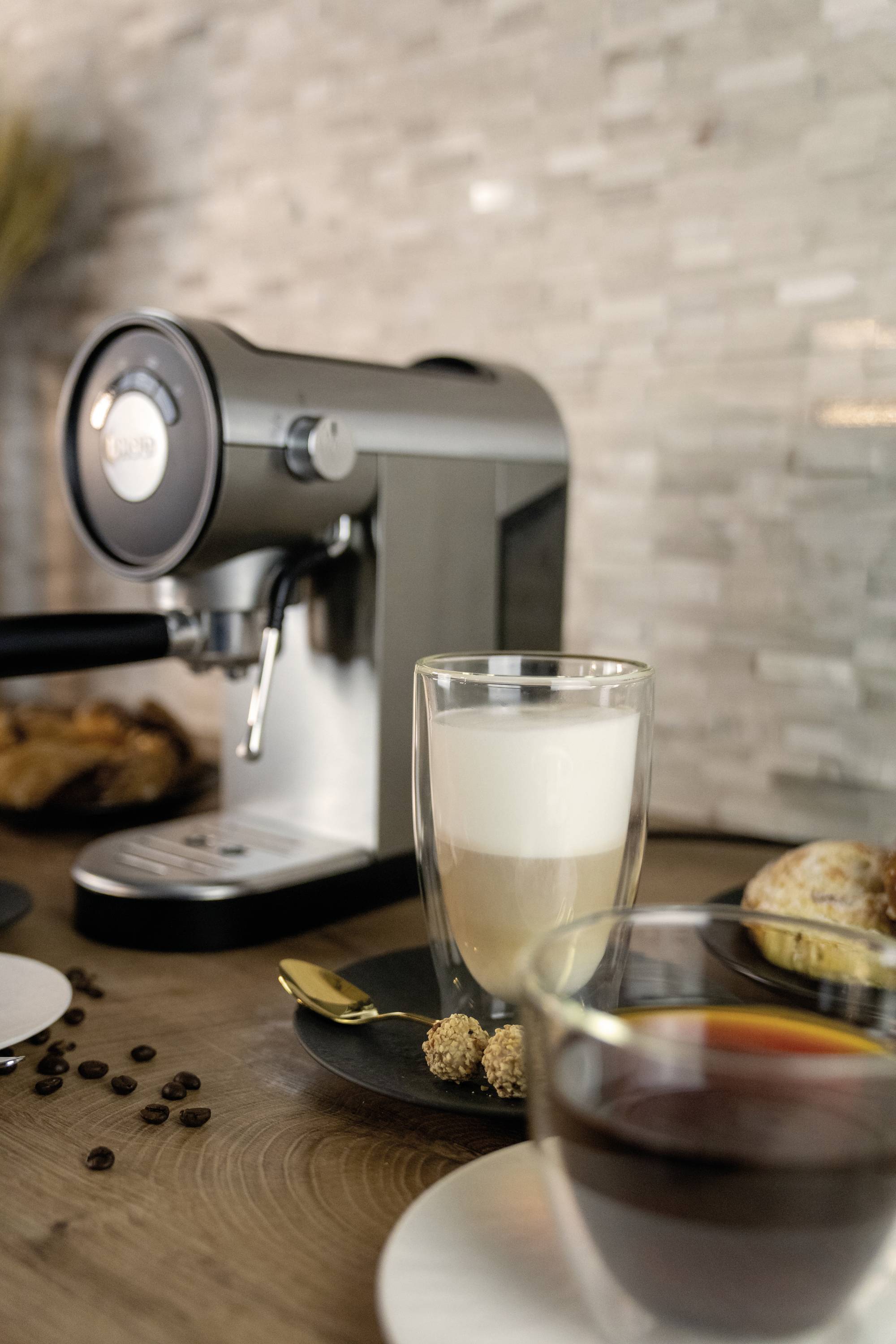 Kaffeemaschine mit einer Tasse Cappuccino im Vordergrund, umgeben von Croissants und Kaffeebohnen auf einem Holztisch.