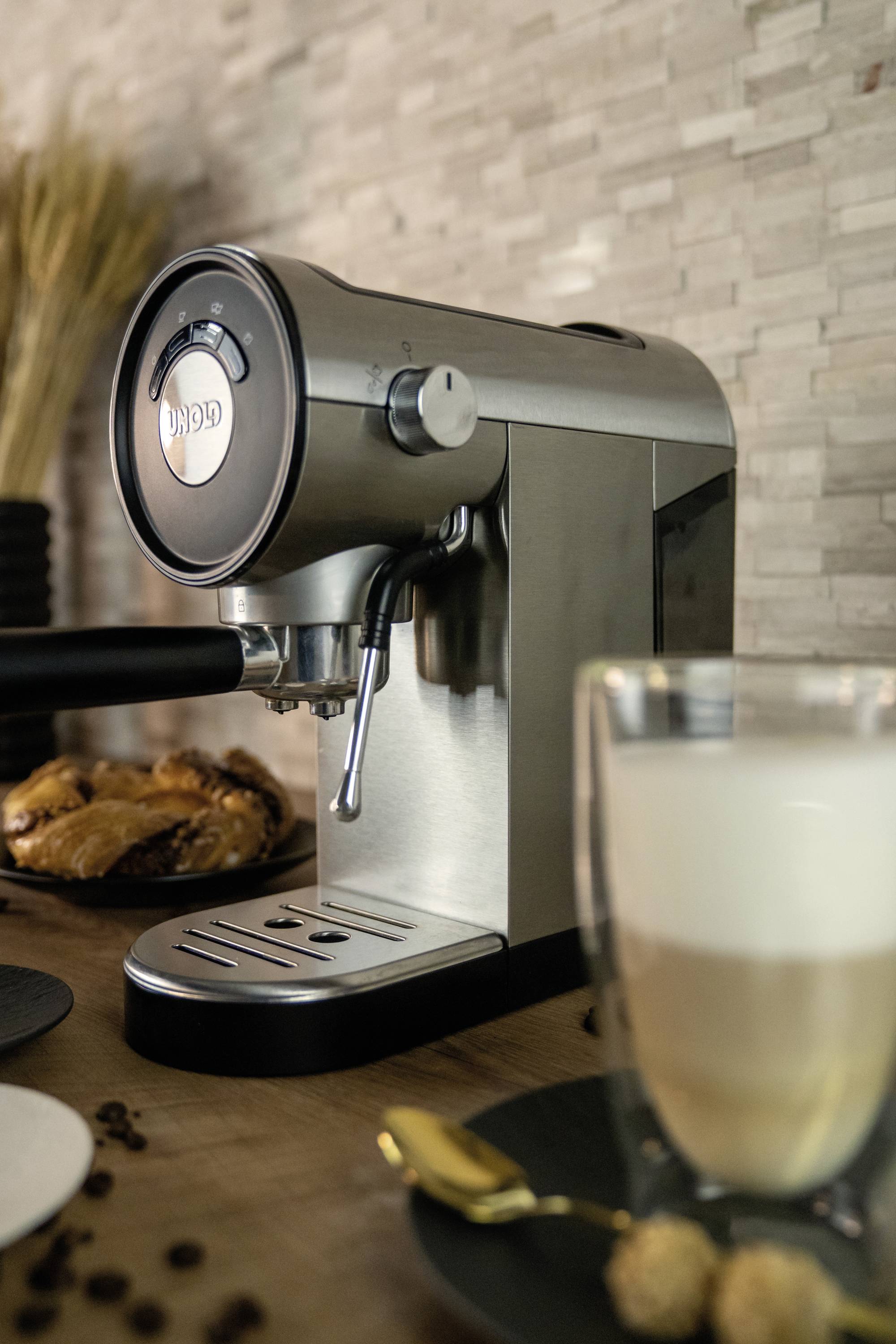 Silberne Espressomaschine mit Latte-Macchiato-Glas und Gebäck auf Holztisch, körnige Wandverkleidung im Hintergrund.