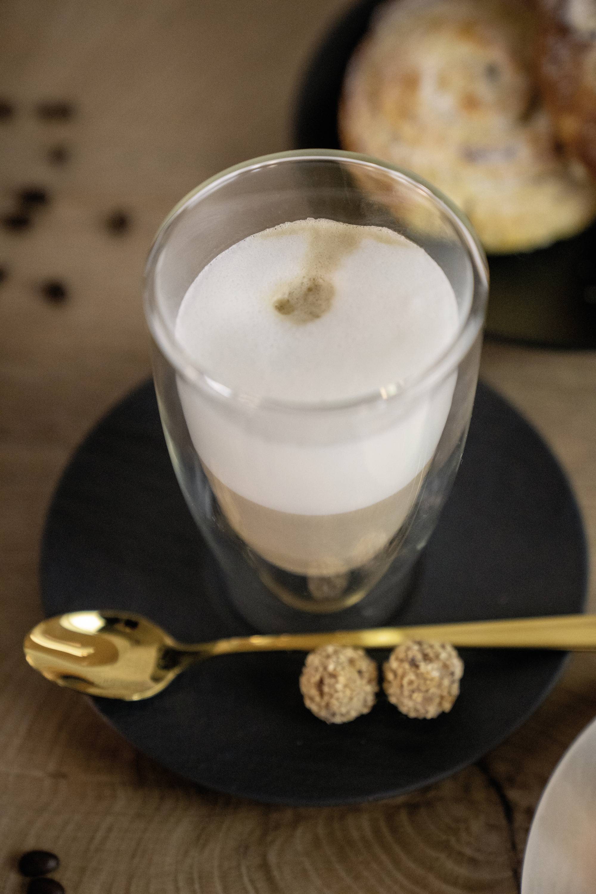 Ein Glas Latte Macchiato auf einem schwarzen Teller mit einem goldenen Löffel und zwei kleinen Keksen. Im Hintergrund ein Gebäck.