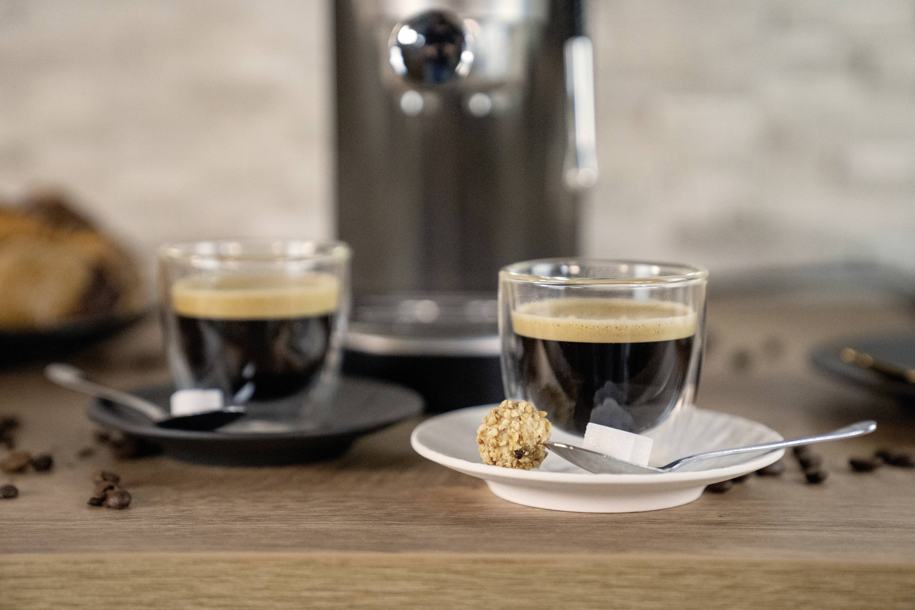 Zwei Espressotassen auf Untertassen mit Löffeln, eine pralinenähnliche Süßigkeit auf einem Löffel. Hintergrund: Kaffeemaschine.