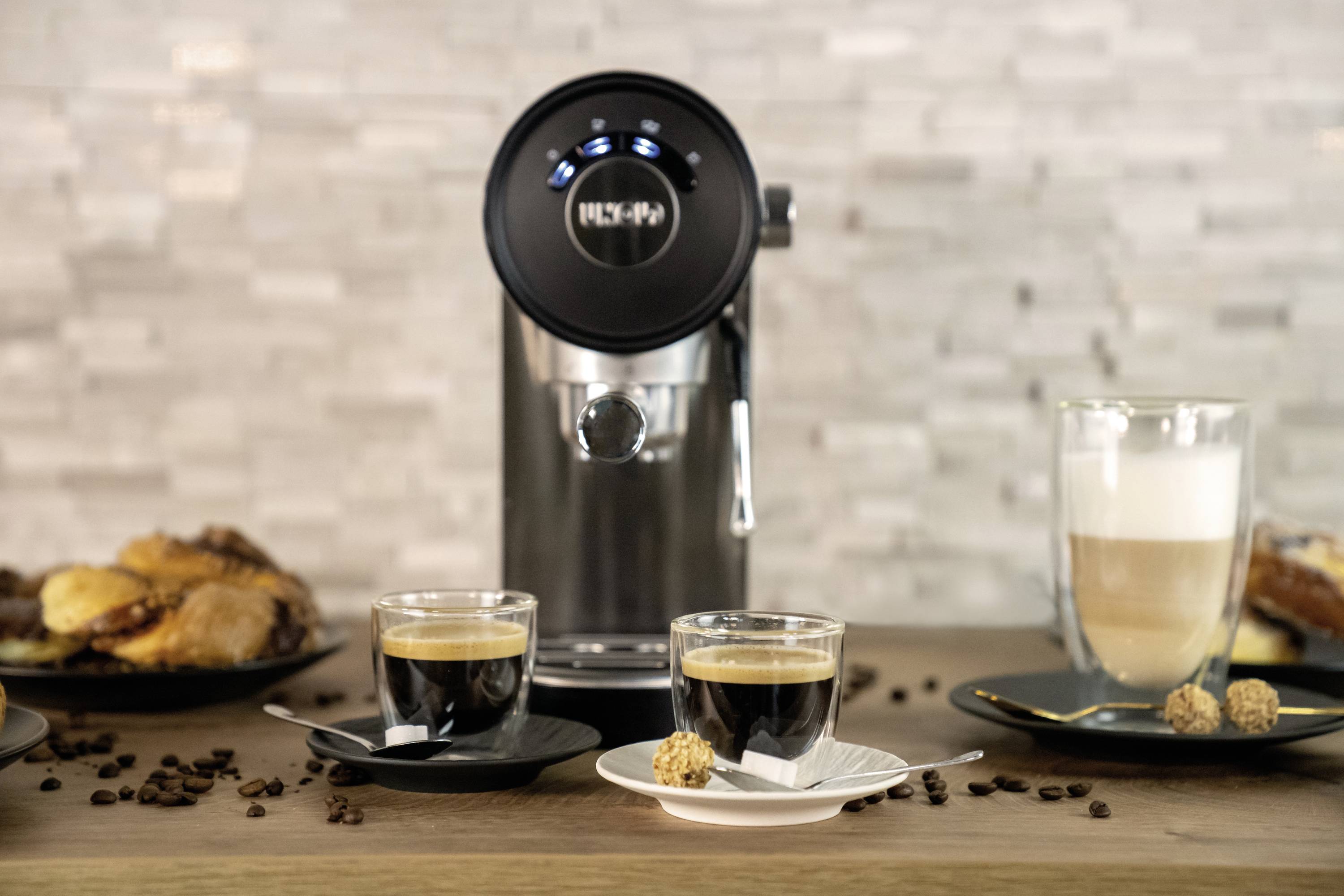 Kaffeemaschine mit zwei Espresso in Gläsern, daneben ein Latte Macchiato, im Hintergrund Croissants und Kaffeebohnen auf einem Tisch.