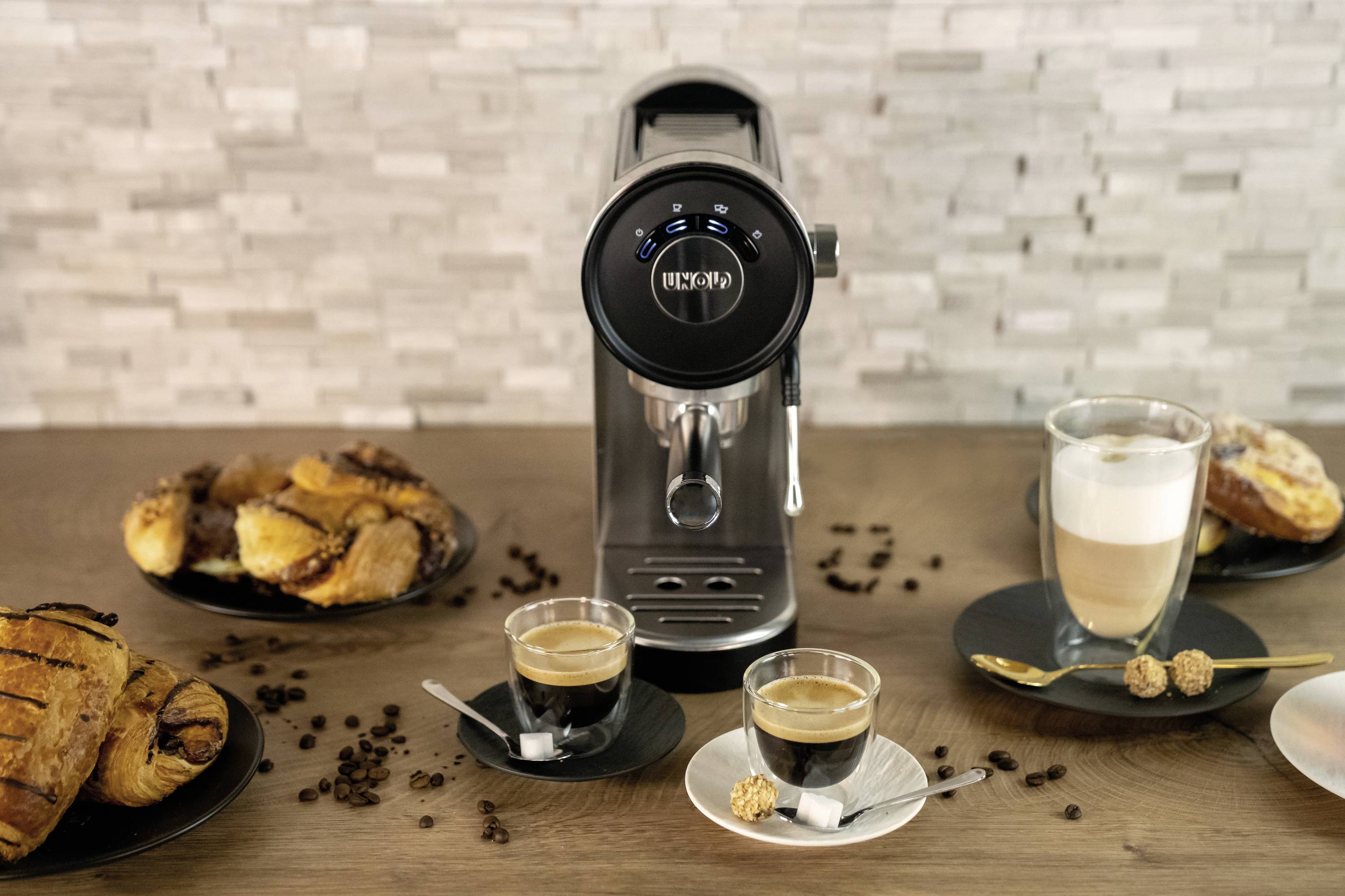 Eine Espressomaschine mit zwei gefüllten Tassen Kaffee auf einem Tisch, umgeben von Gebäck und Kaffeebohnen, vor einer Mosaikwand.