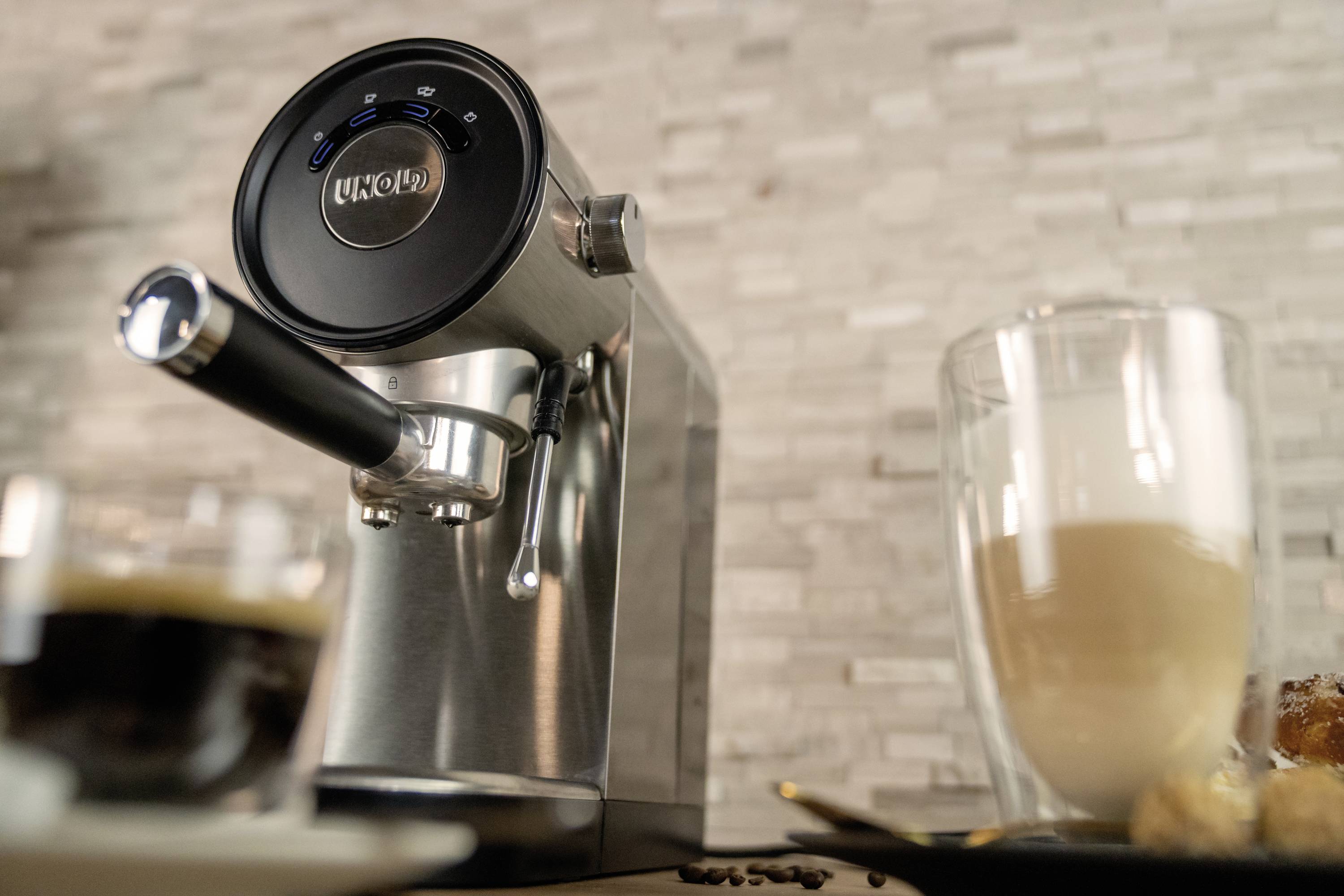 Eine silberne Espressomaschine mit schwarzem Bedienfeld, daneben ein halb gefülltes Latte-Glas auf einem Tablett. Hintergrund aus Steinfliesen.