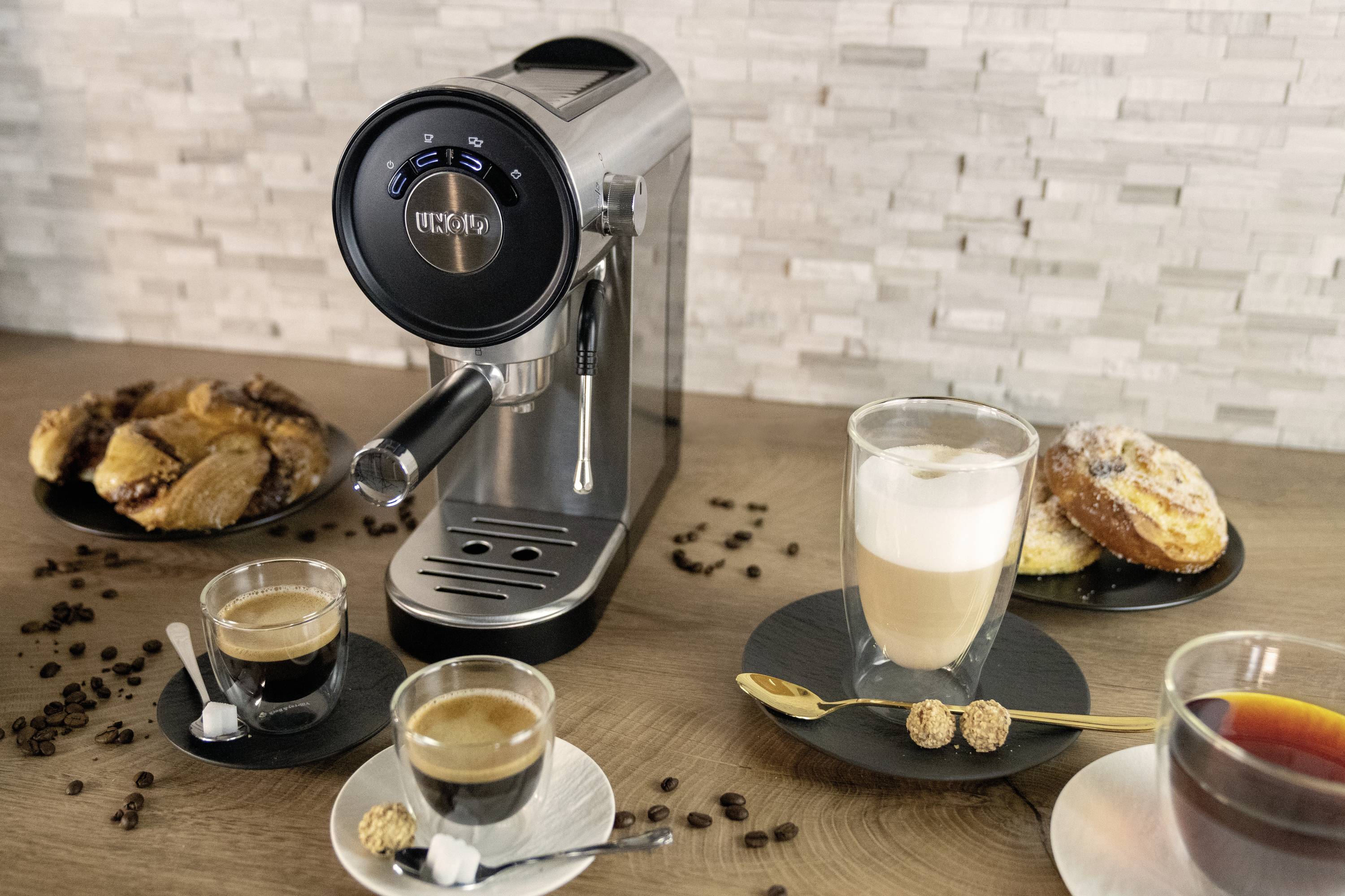 Eine silberne Espressomaschine auf einem Tisch, umgeben von Tassen mit Espresso und Latte Macchiato, Gebäck und Kaffeebohnen.