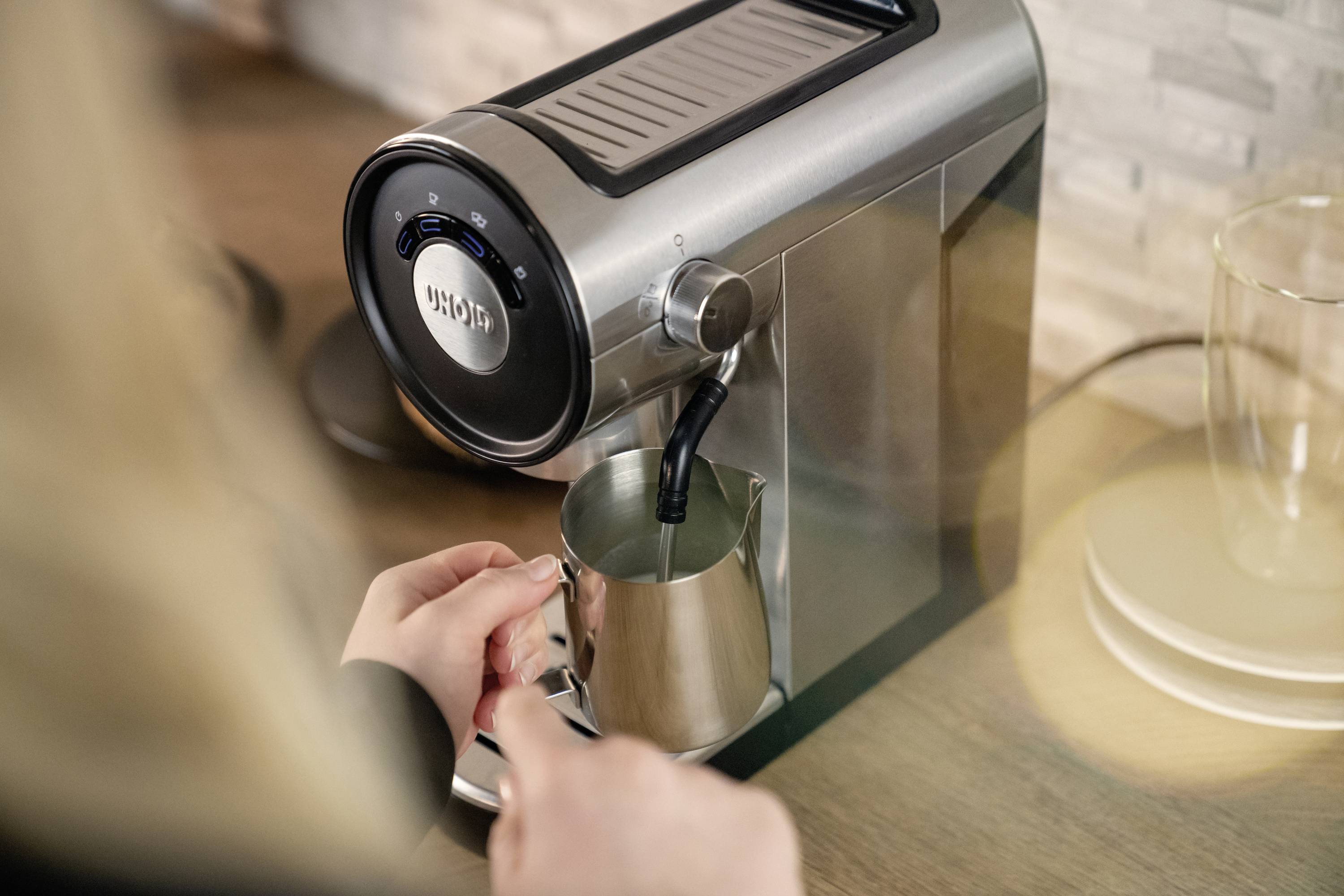Eine Person schäumt Milch mit einer silbernen Kaffeemaschine in einem Edelstahlkrug auf. Im Hintergrund befindet sich eine Küchenarbeitsplatte.