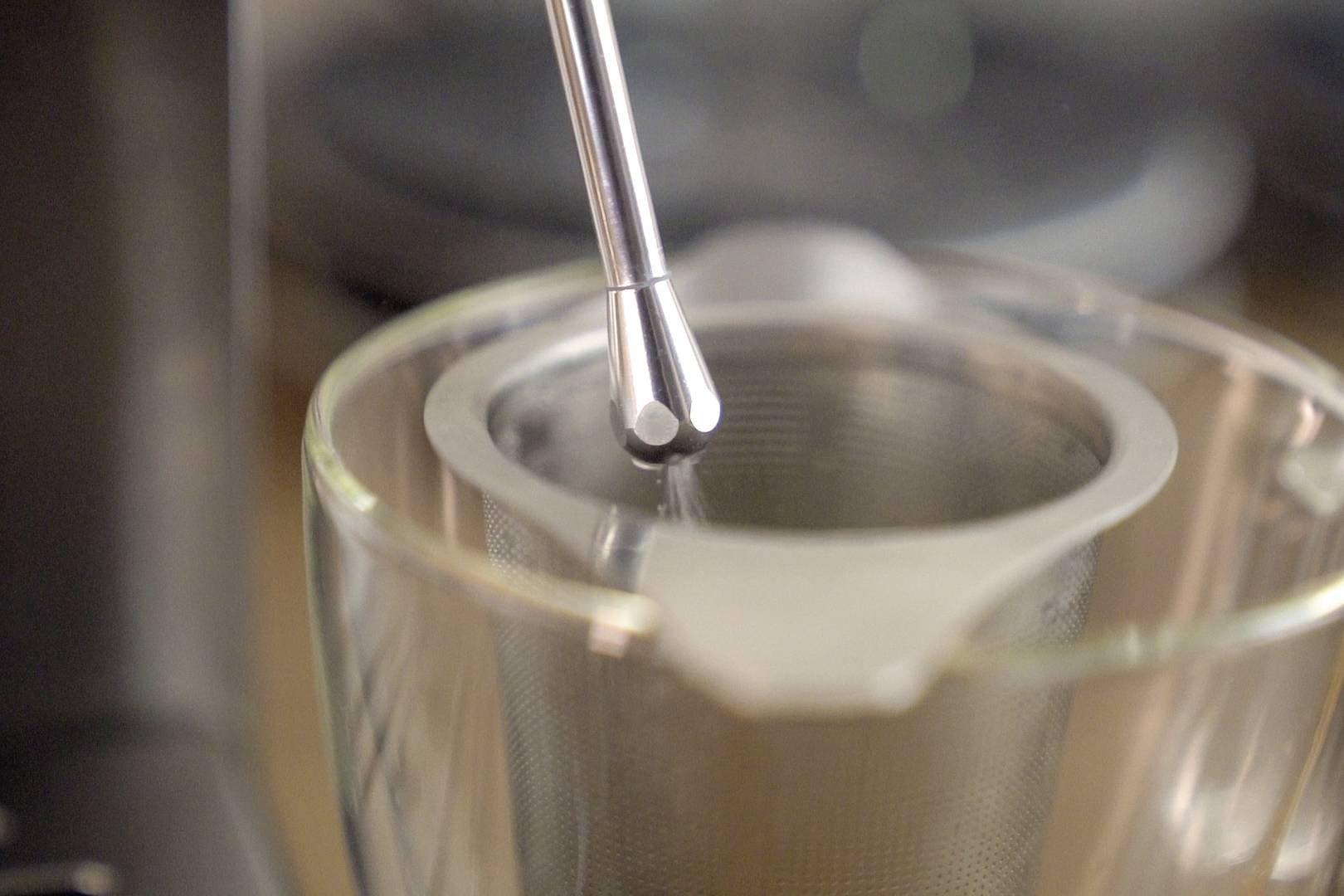 Nahaufnahme eines Teesiebs in einem Glasbecher, darauf ein Metalllöffel. Der Fokus liegt auf der Zubereitung von heißem Tee.
