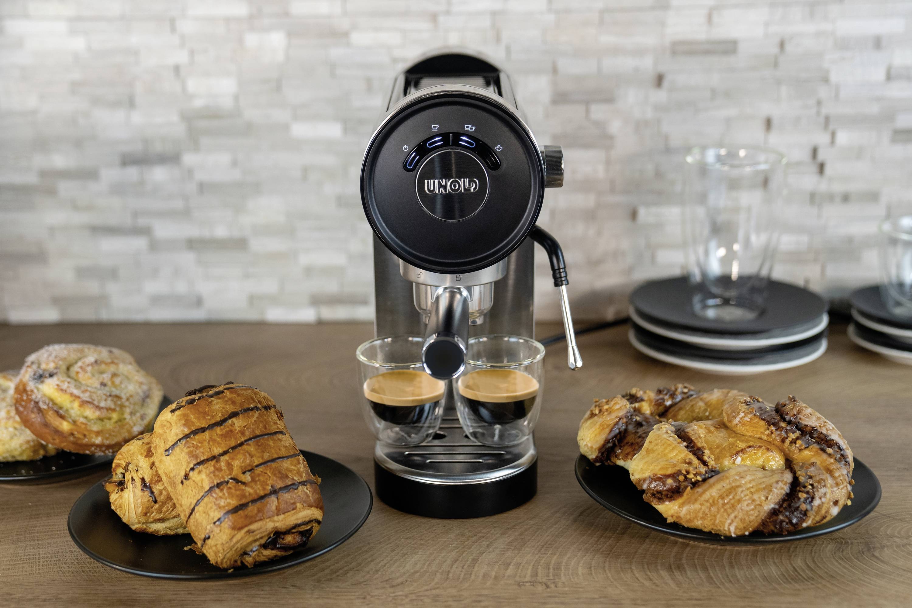 Kaffeemaschine beim Brühen von zwei Espressi, flankiert von verschiedenen Gebäckstücken auf Tellern; moderne Küche im Hintergrund.