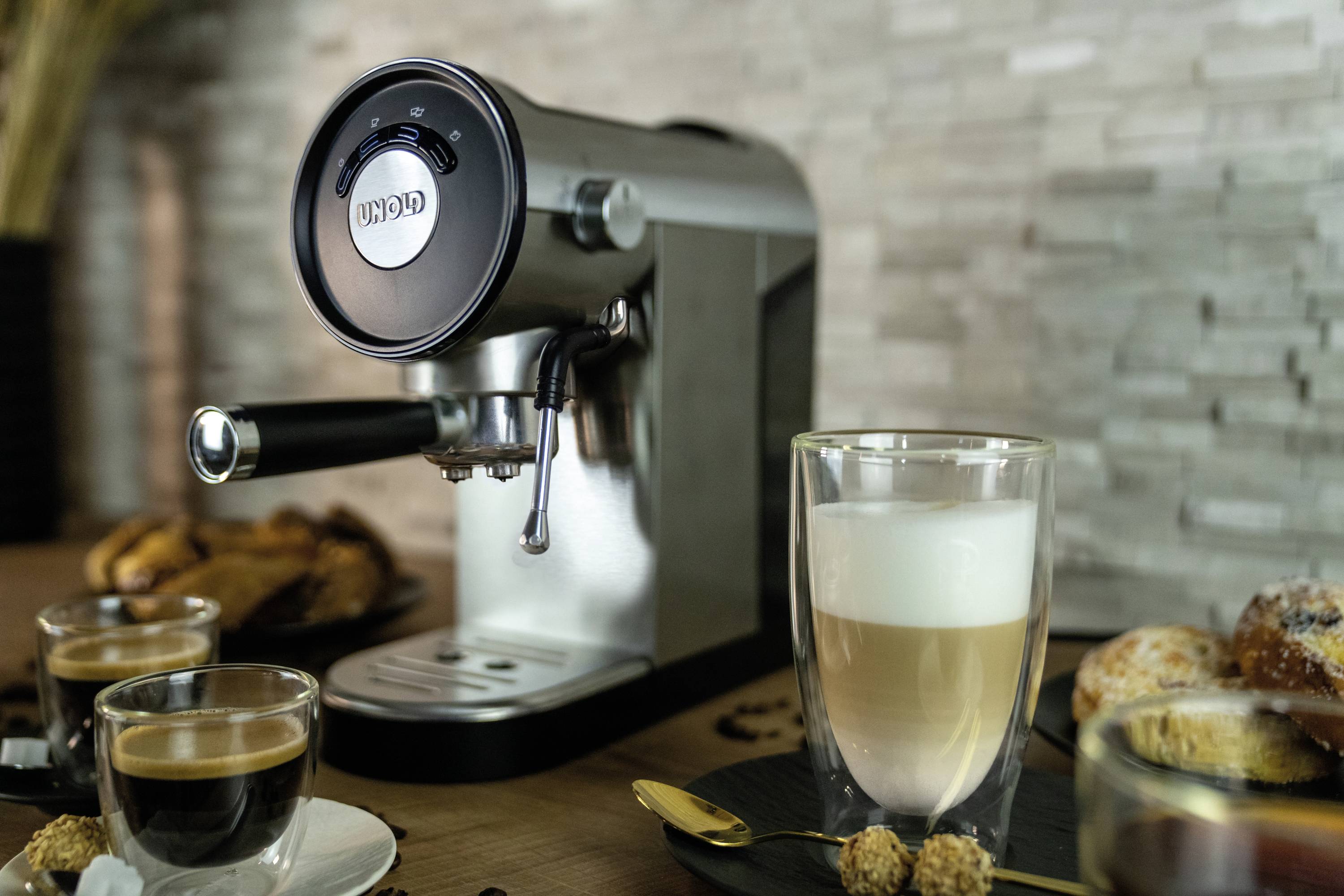 Eine silberne Espressomaschine auf einem Holztisch, umgeben von zwei Tassen Kaffee und Gebäck vor einer Steinwand.