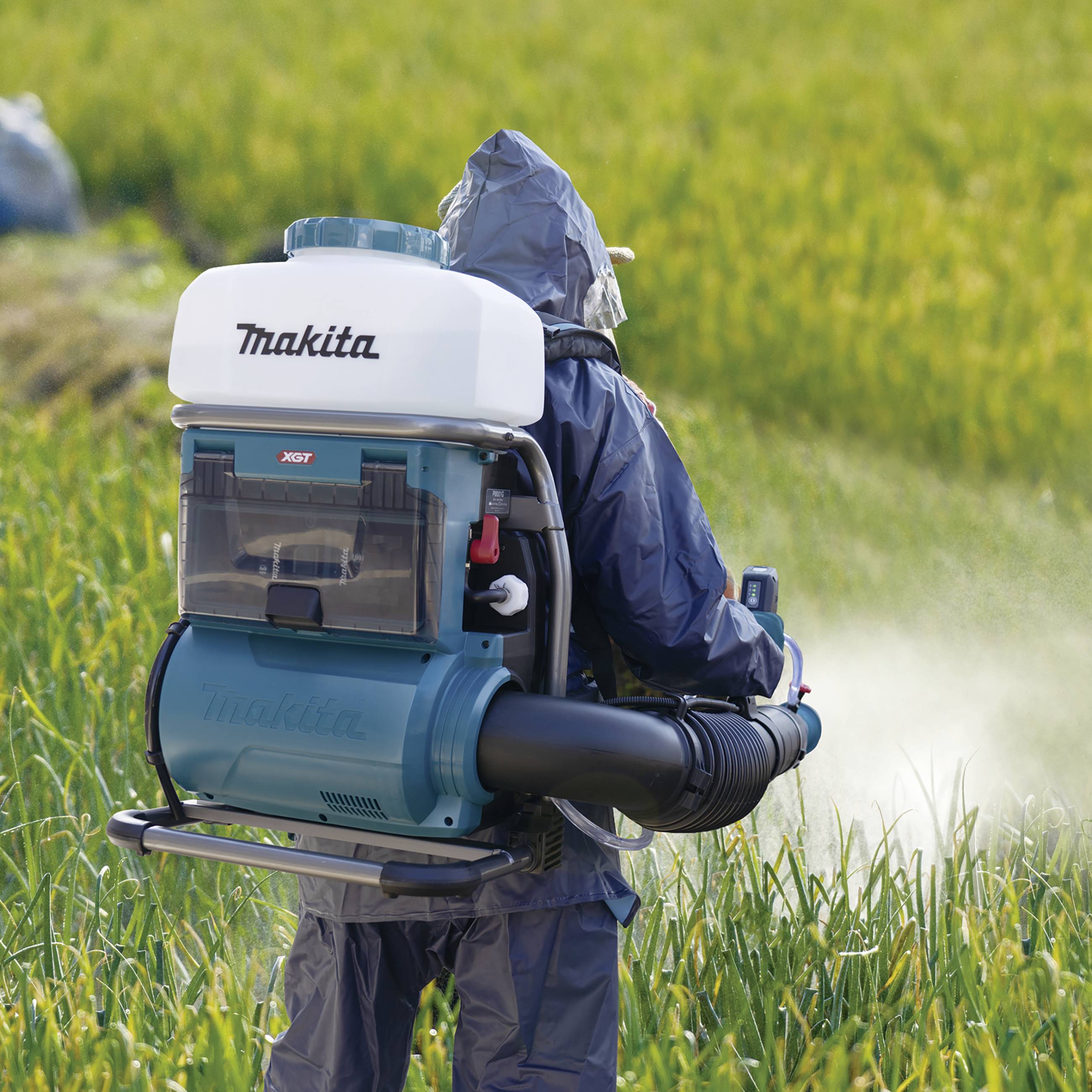 Eine Person in Schutzkleidung verwendet im Freien ein tragbares Sprühgerät, um Pflanzen auf einem grünen Feld zu behandeln.