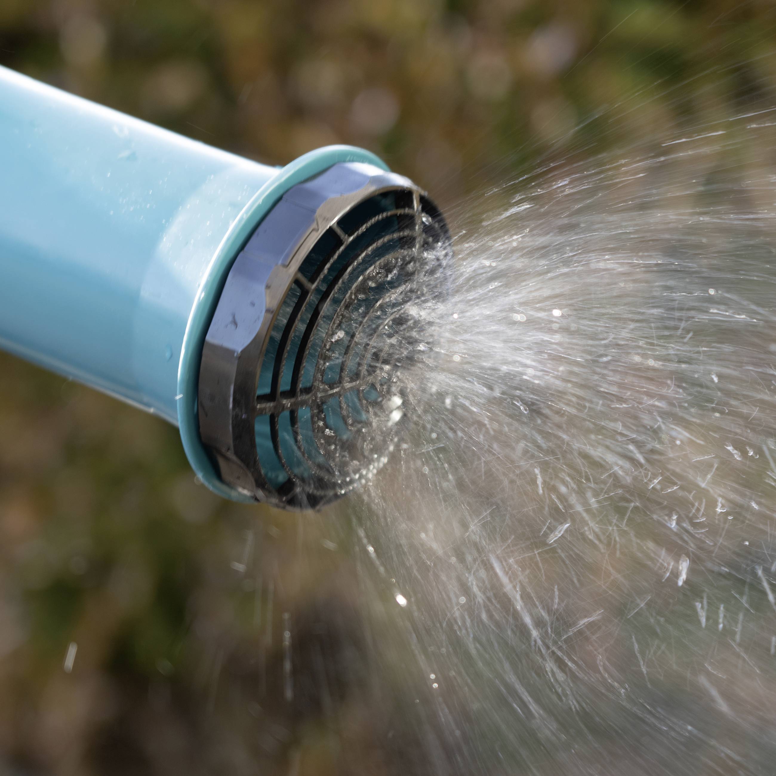Ein blauer Gartenschlauch spritzt einen Wasserstrahl in die Luft vor einem unscharfen Hintergrund, vermutlich im Freien.