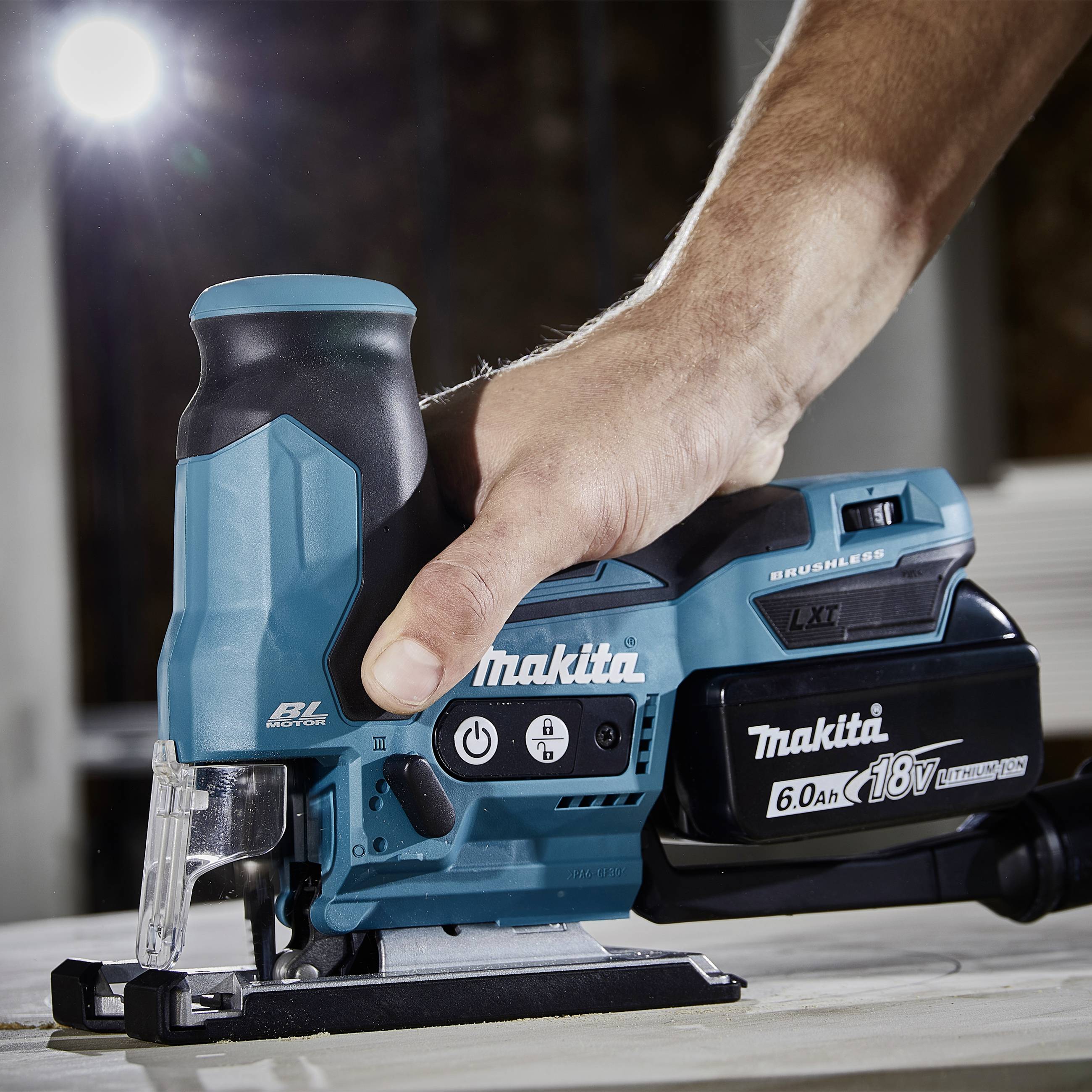 Eine Hand hält eine elektrische Stichsäge der Marke Makita, die auf einem Holztisch liegt. Die Säge ist in Benutzung.