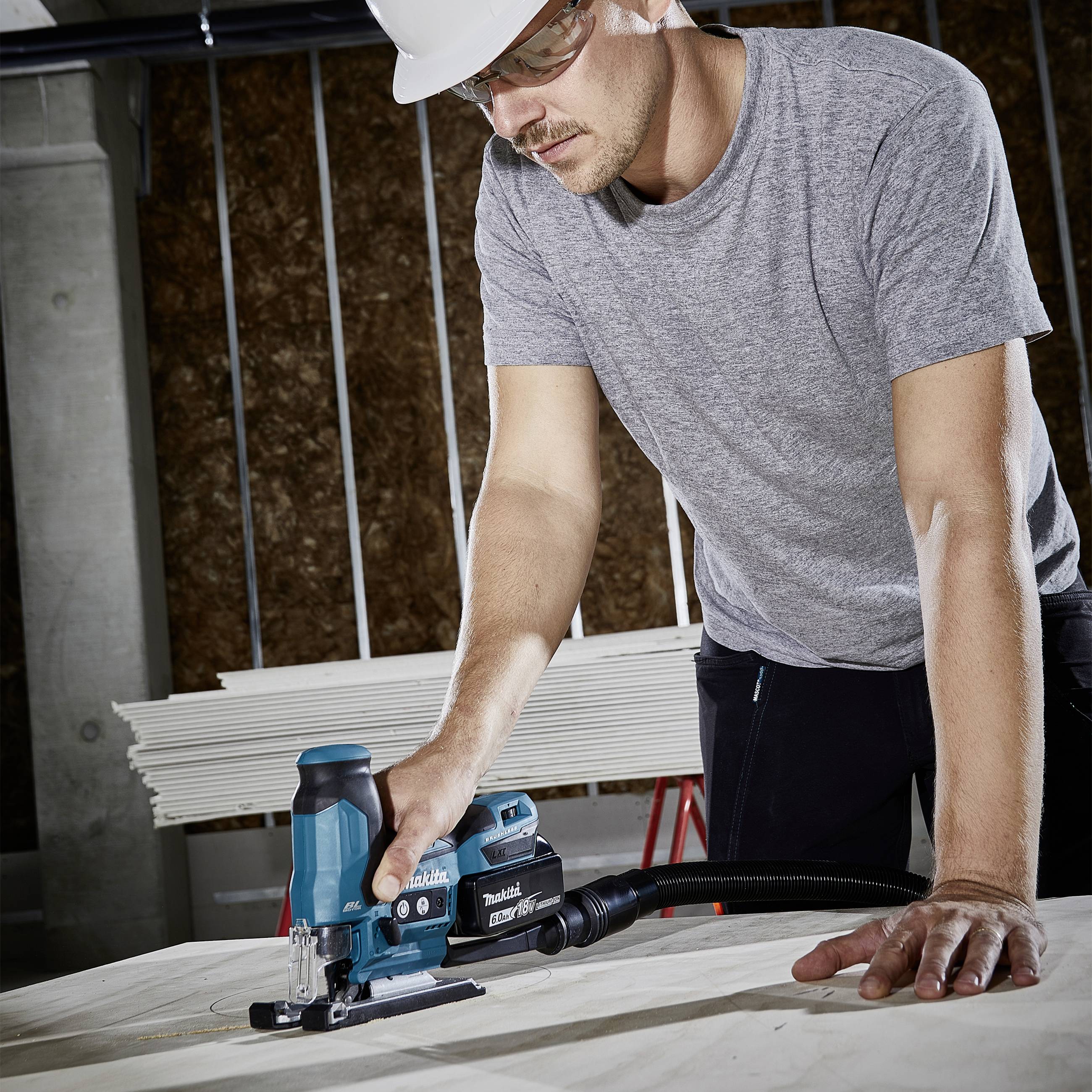 Ein Mann mit Schutzbrille und Helm arbeitet mit einer elektrischen Stichsäge an einem Holzbrett in einer Werkstatt.