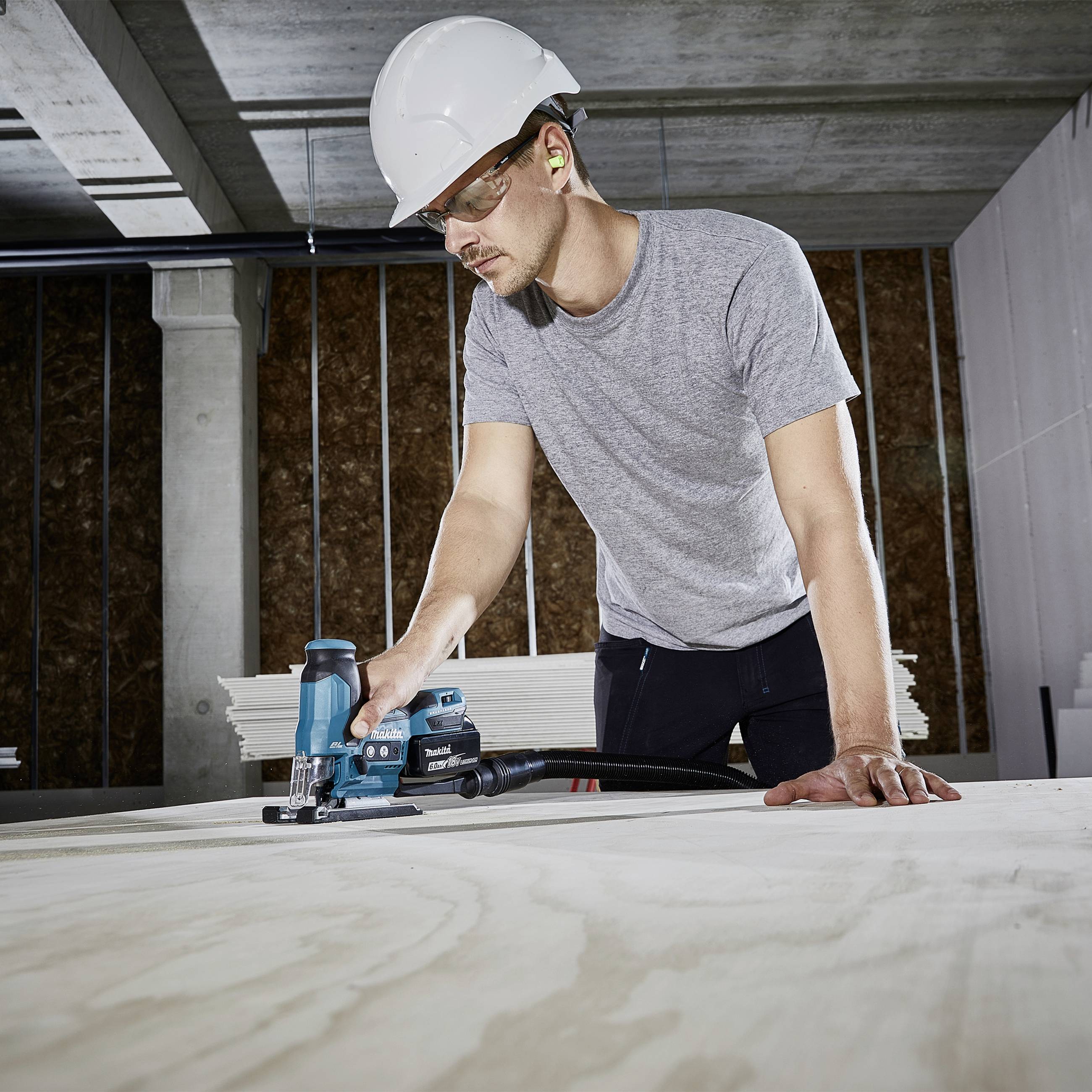 Ein Arbeiter mit Schutzhelm und Gehörschutz benutzt ein Elektrowerkzeug, um Sperrholz in einer Baustelle zu schneiden.