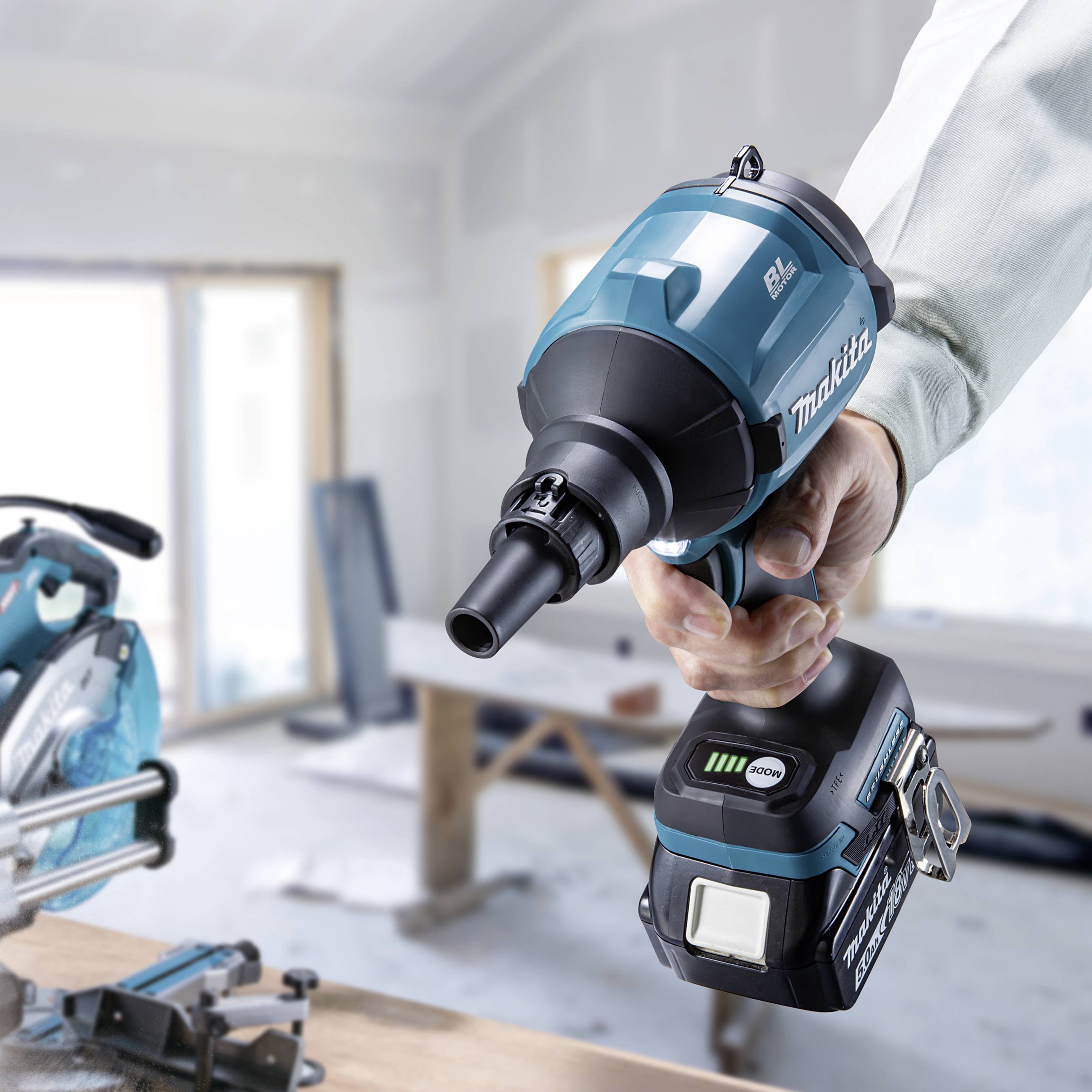 Eine Person hält einen blauen Makita-Akkuschlagschrauber in einer Werkstatt. Im Hintergrund sind unscharfe Werkzeuge zu sehen.