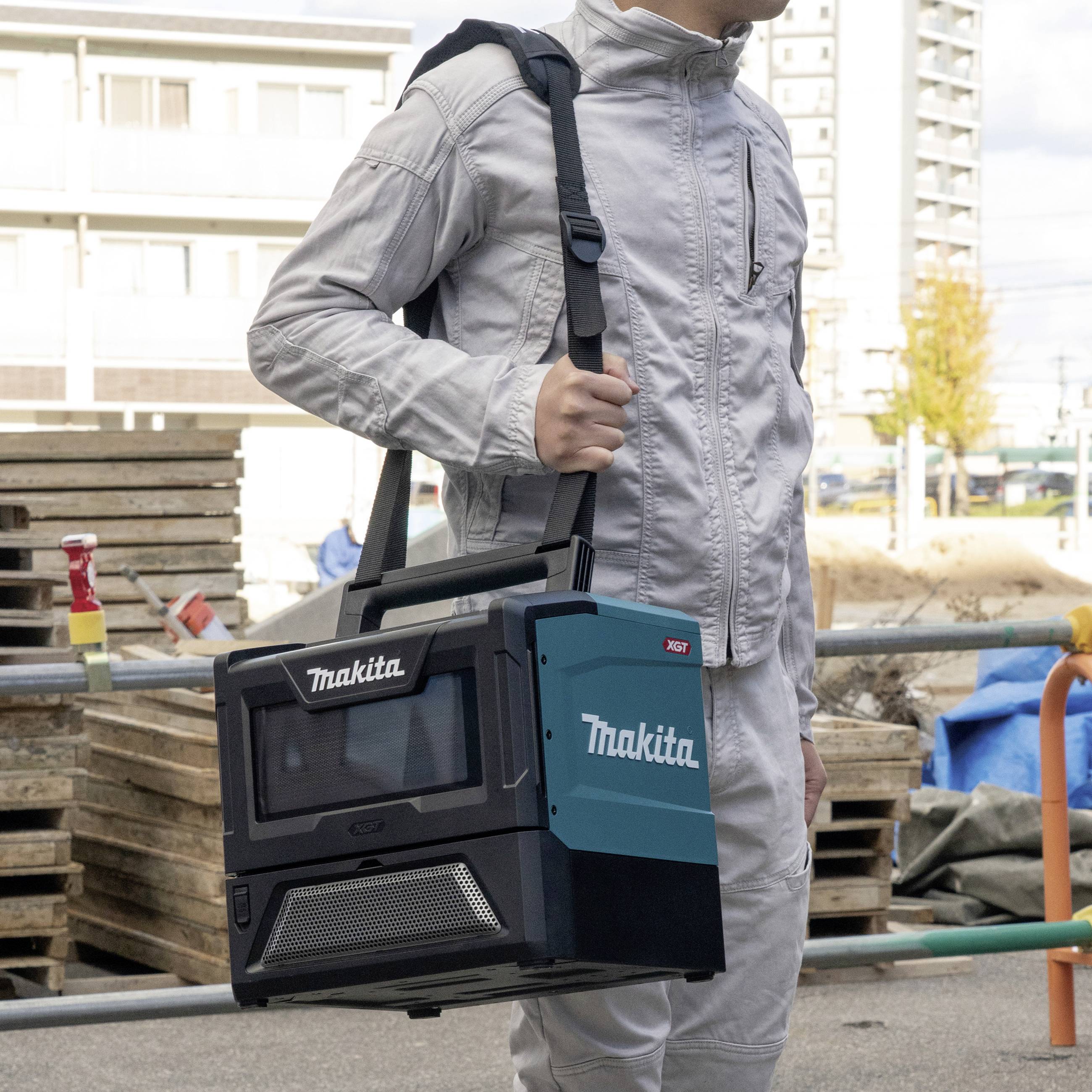 Eine Person trägt einen tragbaren, akkubetriebenen Makita-Heizlüfter auf einer Baustelle. Im Hintergrund sind Baugeräte sichtbar.
