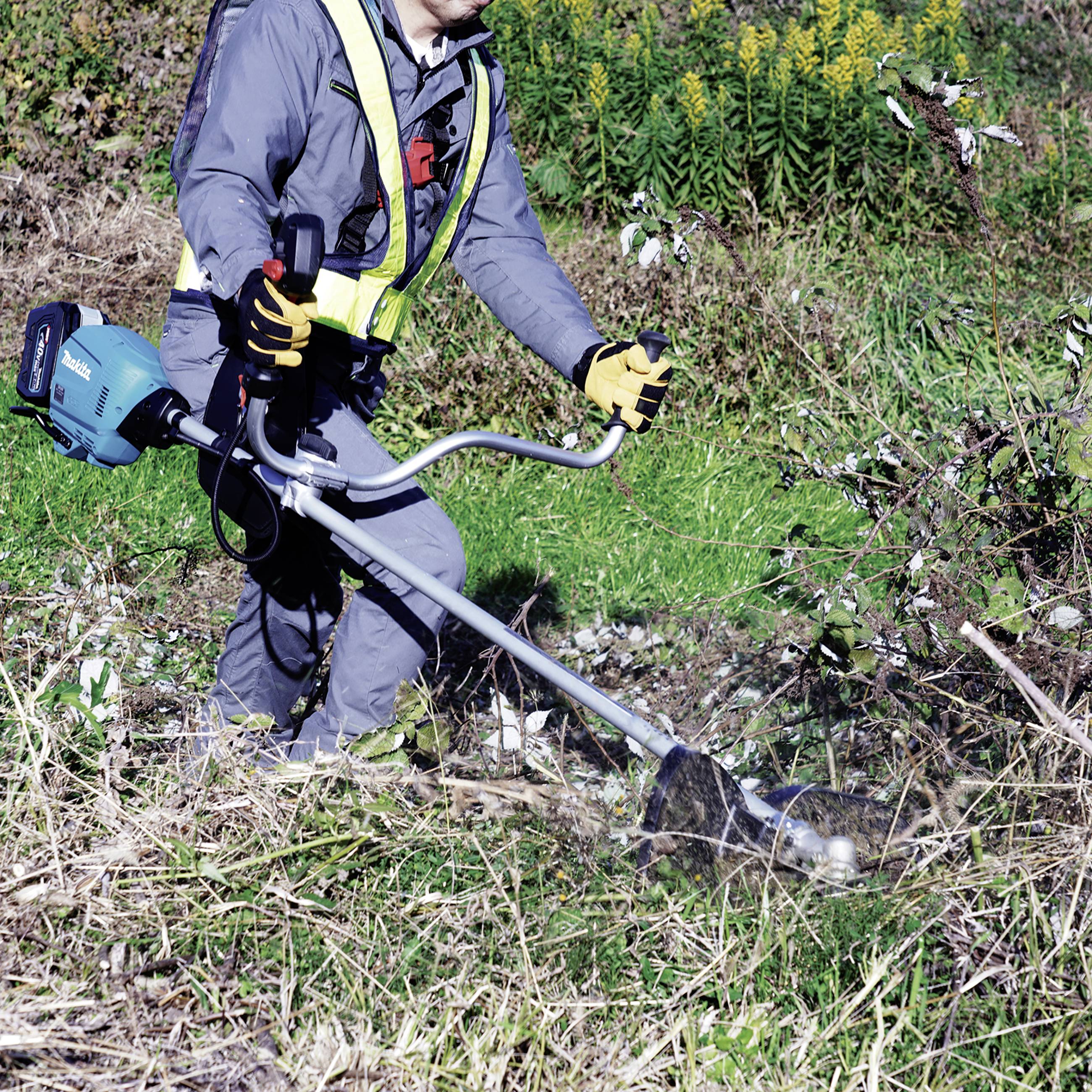 Eine Person in Arbeitskleidung benutzt einen Freischneider, um Gras und Büsche in einem bewachsenen Bereich zu trimmen.