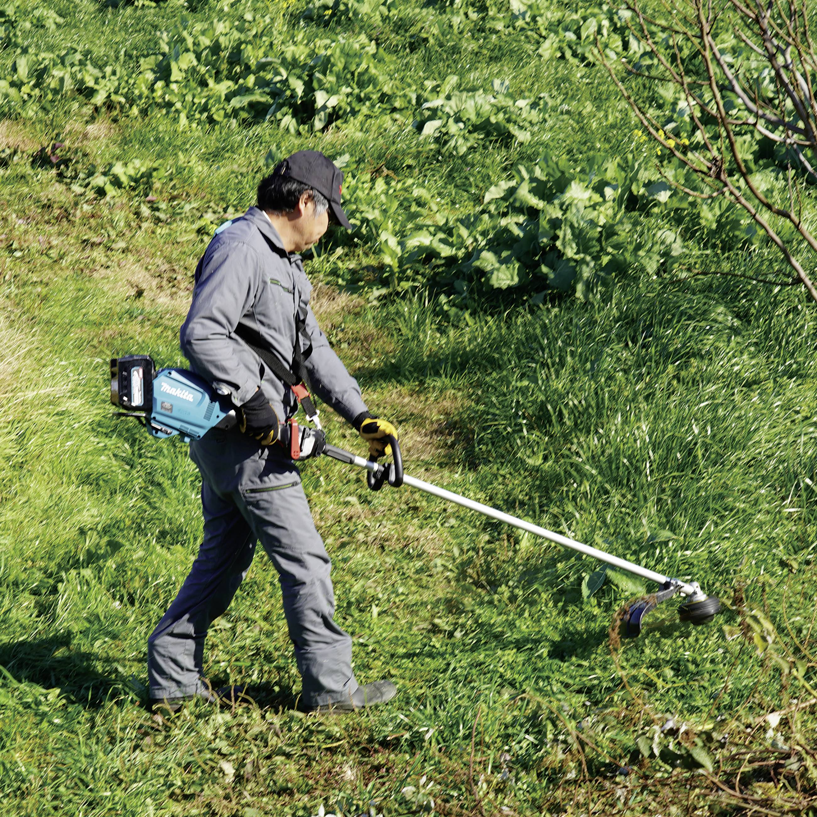 Eine Person in Arbeitskleidung schneidet mit einem Rasentrimmer Gras auf einer grünen Wiese. Im Hintergrund sind Büsche zu sehen.
