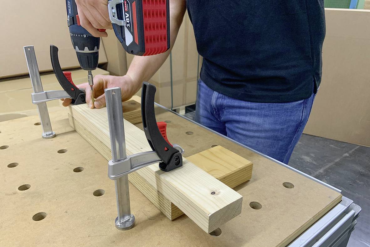 Eine Person fixiert ein Holzstück auf einem Tisch mit Klemmen und bohrt ein Loch.