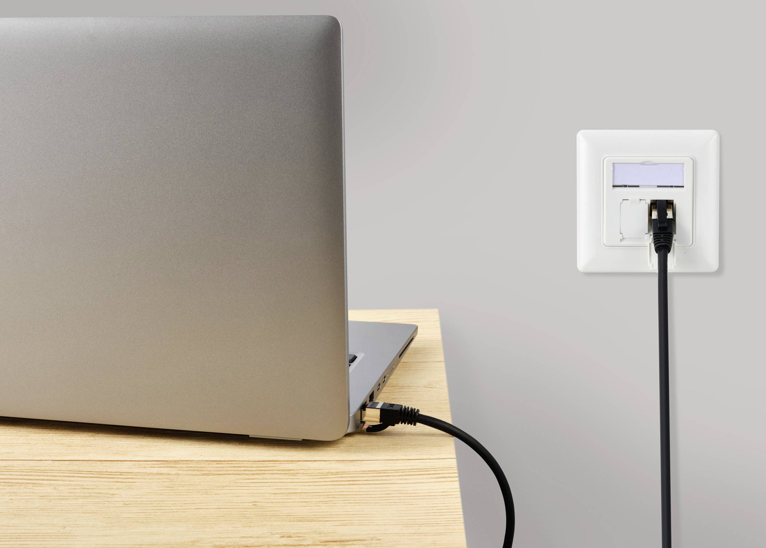 Ein Laptop auf einem Holztisch wird über ein schwarzes Ladegerät an einer weißen Steckdose an der Wand aufgeladen.