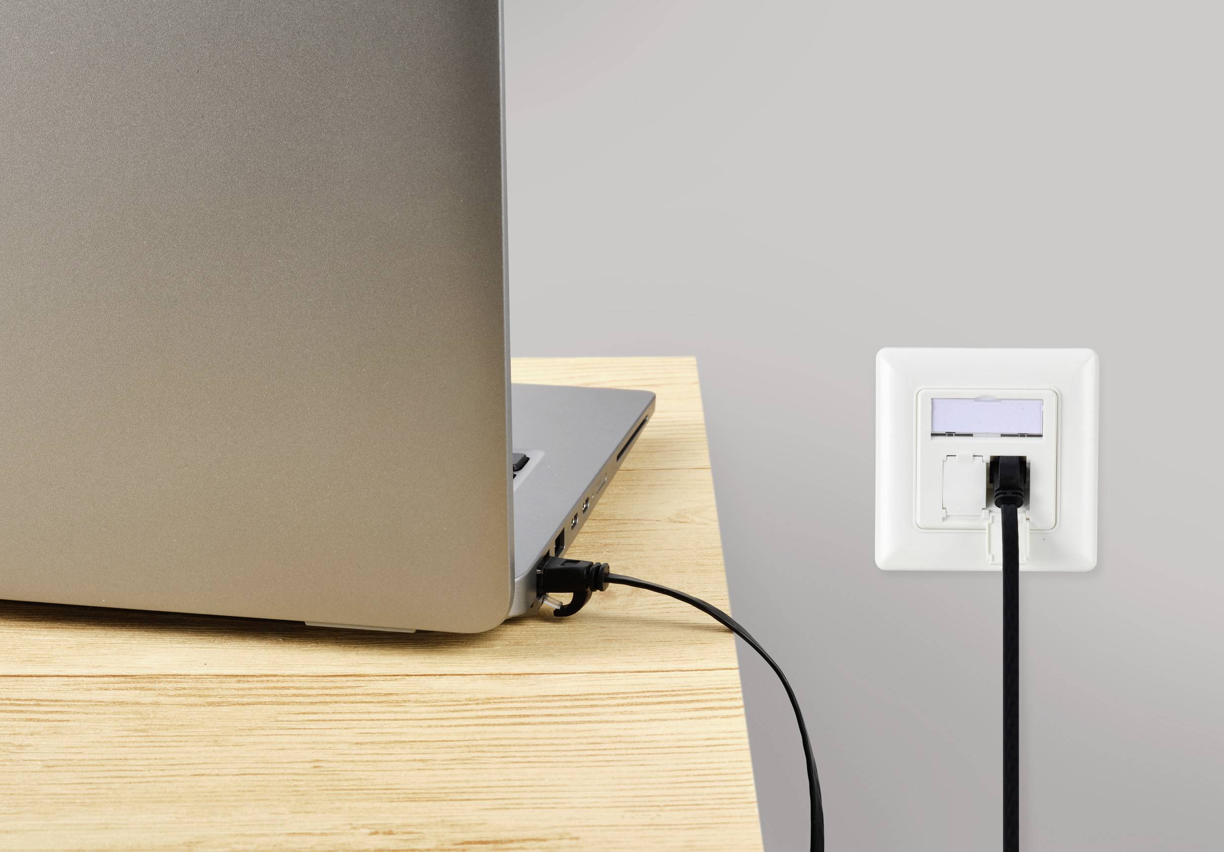 Ein Laptop wird auf einem Holztisch über ein schwarzes Kabel an eine Wandsteckdose angeschlossen.
