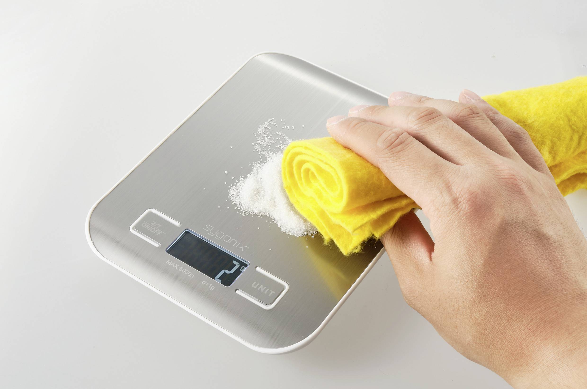 Hand wischt weißen Stoff von digitaler Küchenwaage mit gelbem Tuch; zeigt Reinigungsvorgang und trägt zur Sauberkeit bei.