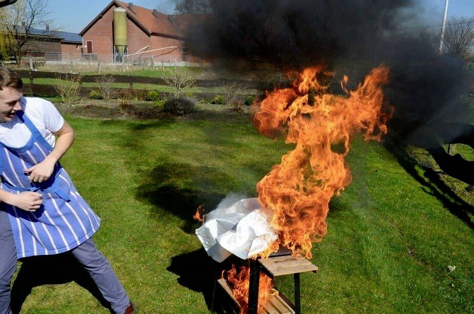 Ein Mann in einem gestreiften Schürze steht in einem Garten und blickt überrascht, während Flammen dramatisch von einem Grill aufsteigen und Rauch darüber wogt.
