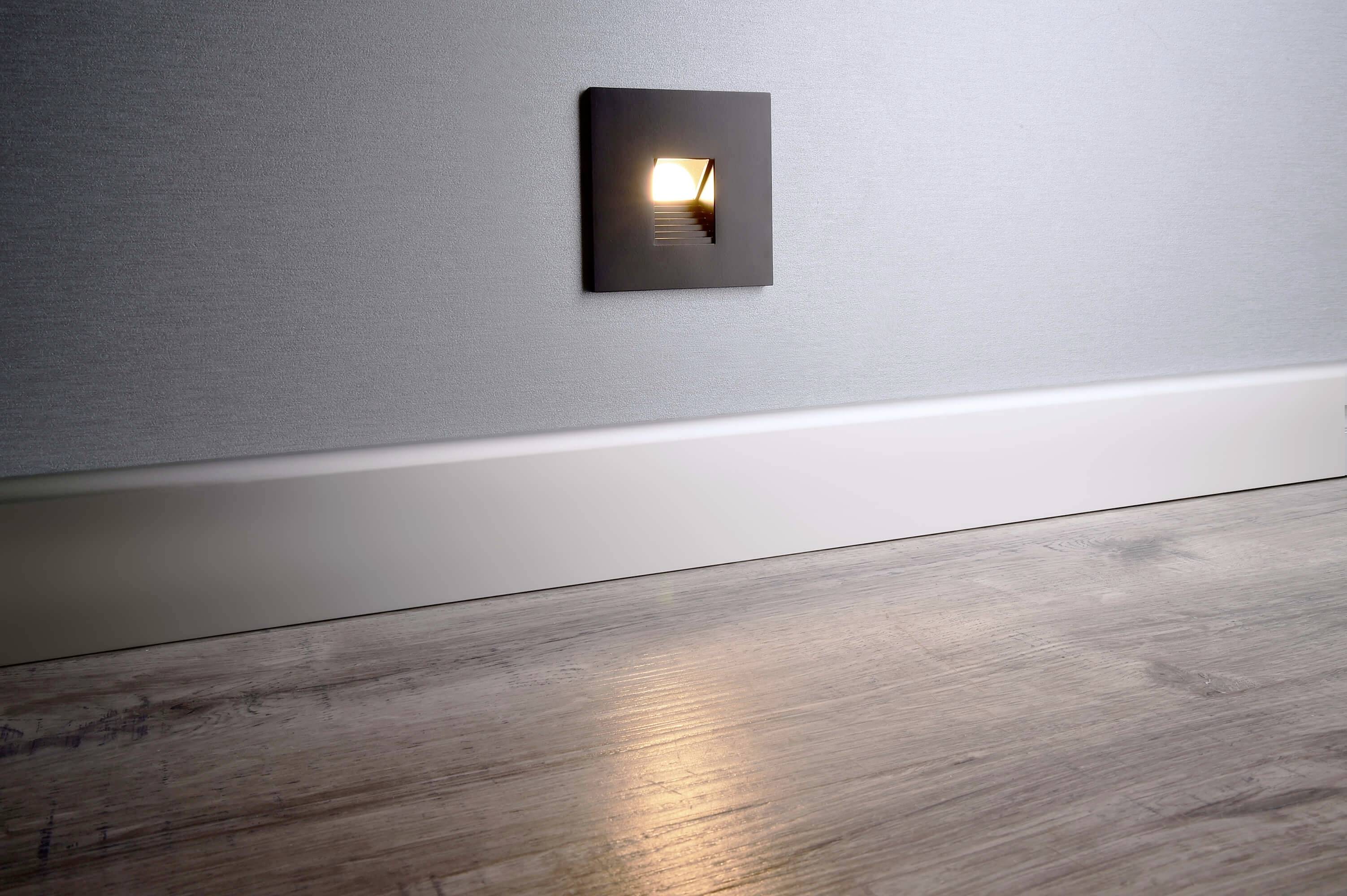 Eine kleine Wandleuchte leuchtet am unteren Teil einer Wand über einem Holzfußboden und erzeugt einen dezenten Lichtschein.