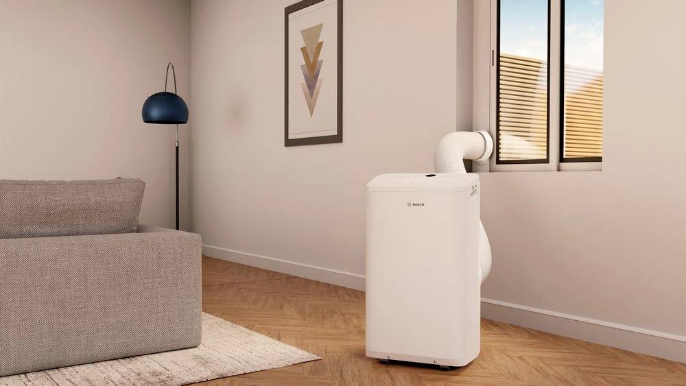Tragbare Klimaanlage in einem modernen Wohnzimmer, verbunden mit einem Fenster. Neben einem grauen Sofa steht eine Stehlampe.